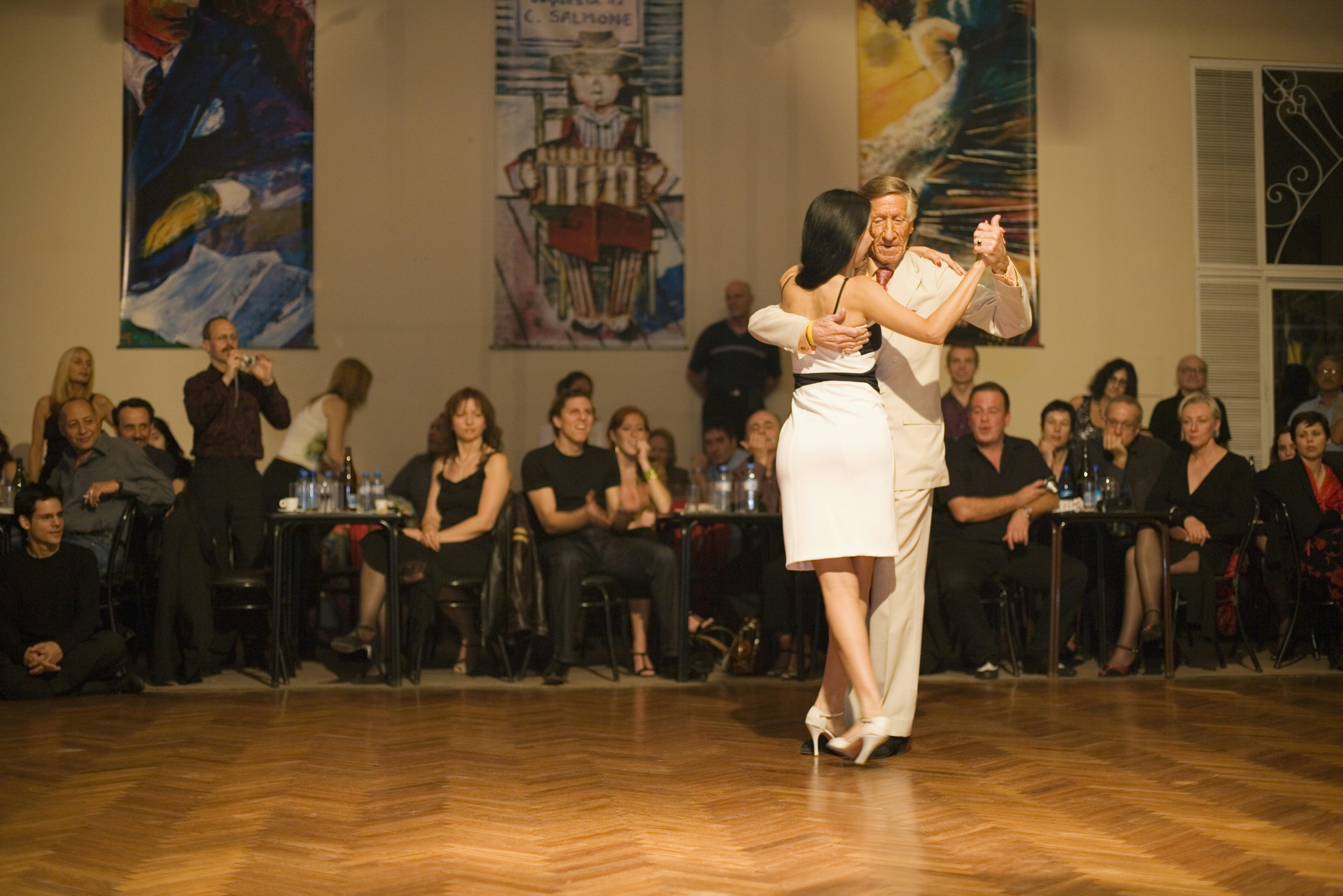 A man in a white suit and a woman in a white dress perform a tango before spectators seated at tables just off the dance floor.