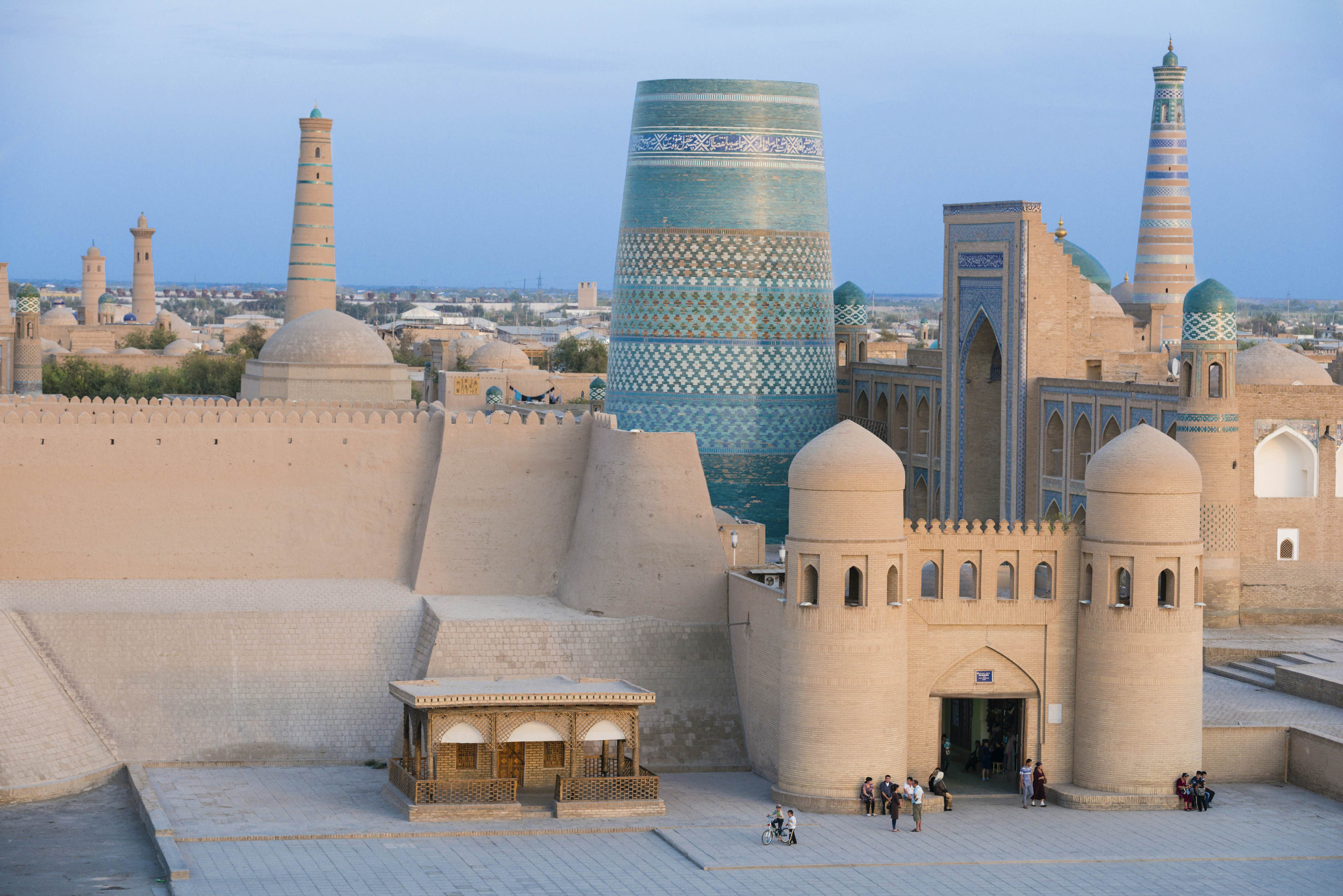 Sand-colored walls with crenellations and a gate with two towers stand in front of a minaret covered in blue tiles. Other minarets and historic buildings are visible in the background.