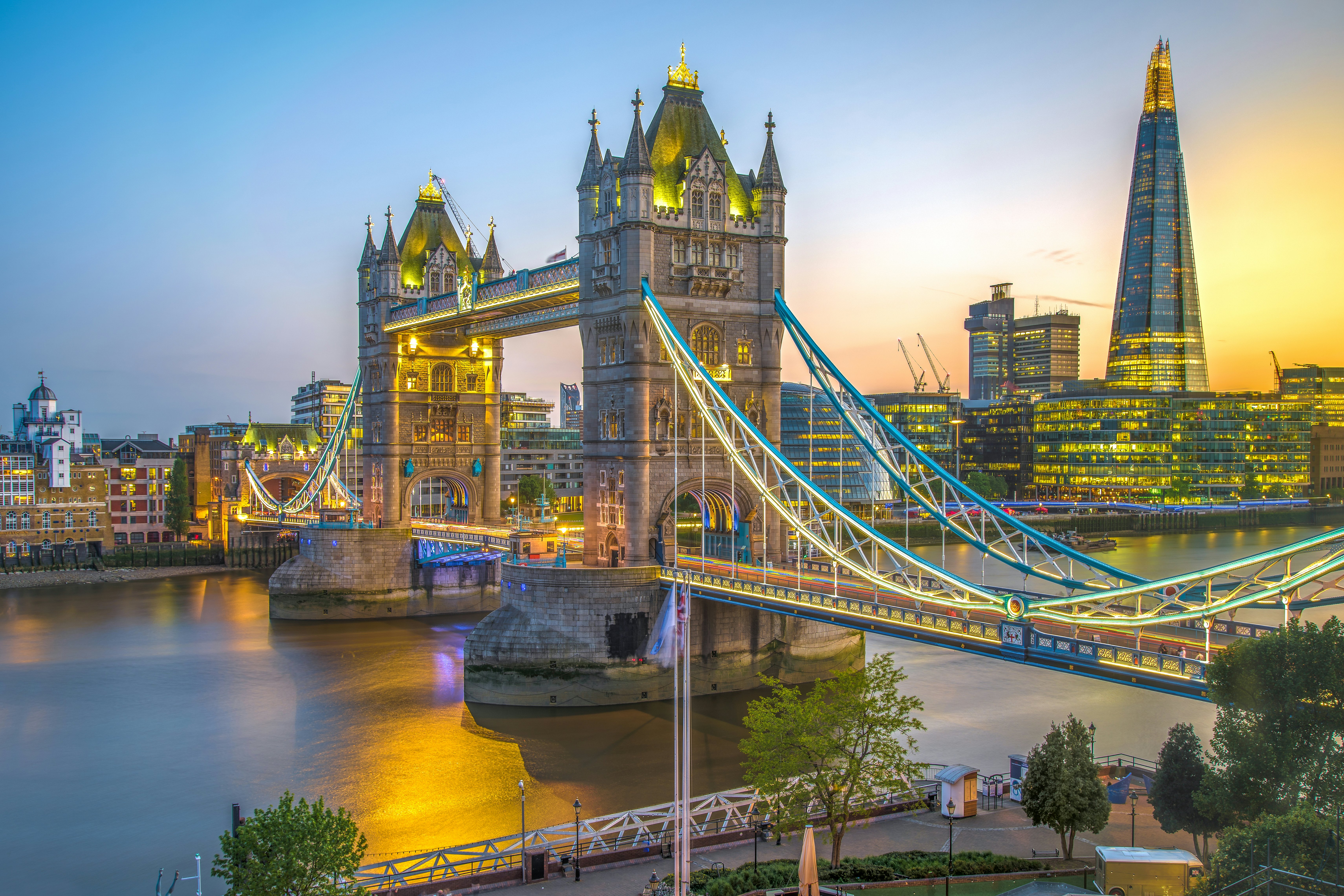 A Victorian bridge with two towers over a river and a large pointed skyscraper at twilight