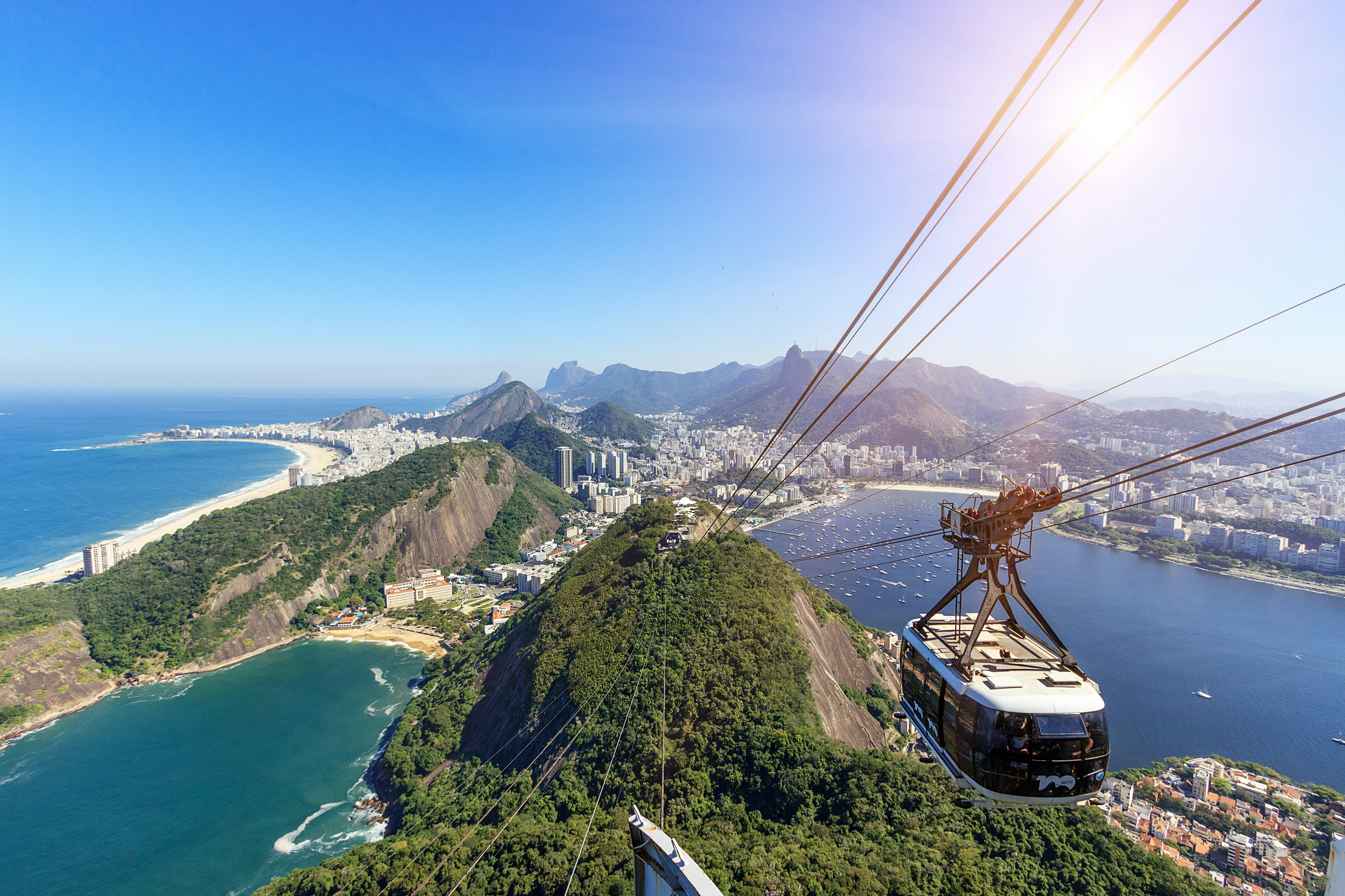 A view looking down at the cable cars ascending a mountain. A hilly, huge city with beaches, bays and apartment towers spreads out below.