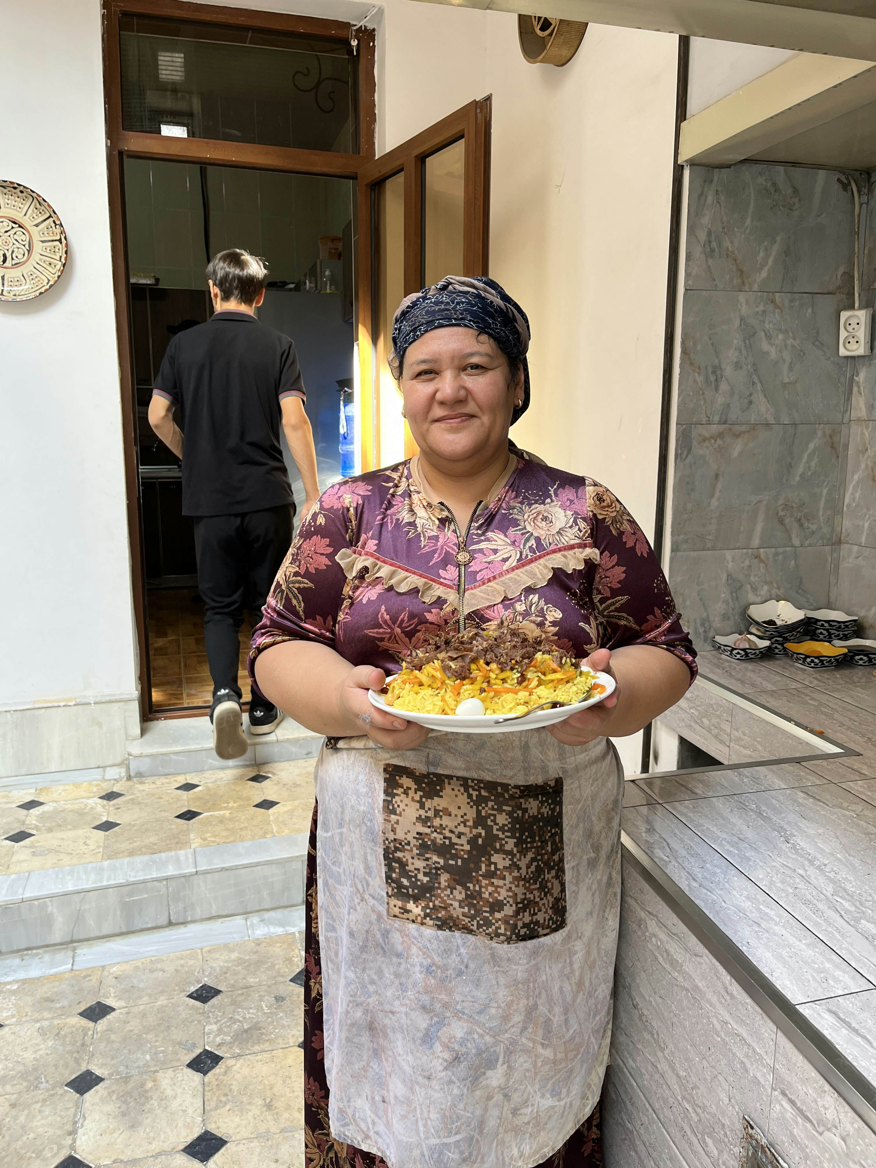 A woman in a head covering stands in kitchen holding a big dish of plov, and Uzbek rice dish.