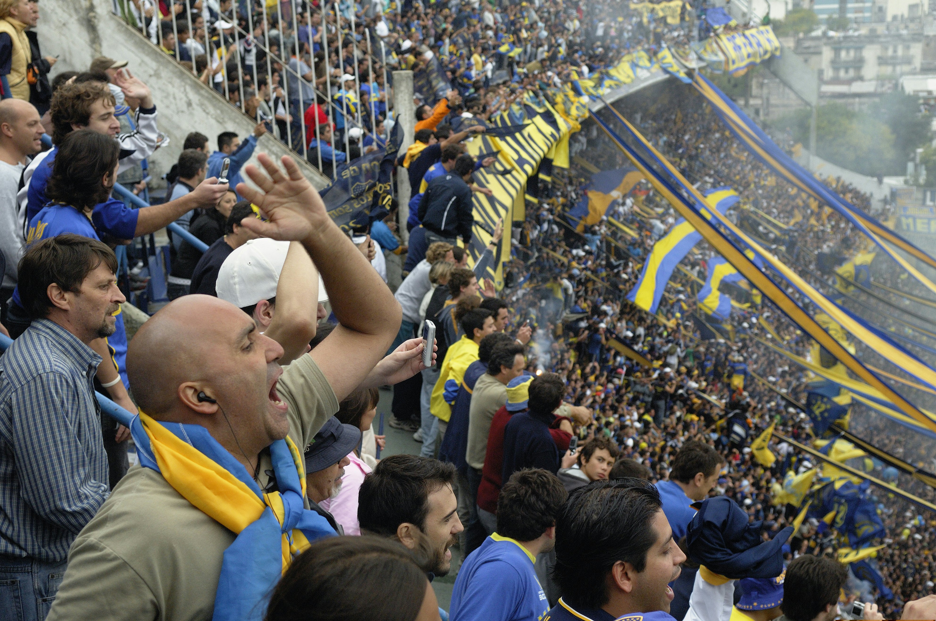 Spectators in home-team colors cheer loudly and wave flags in the bleachers at a match in a packed stadium.