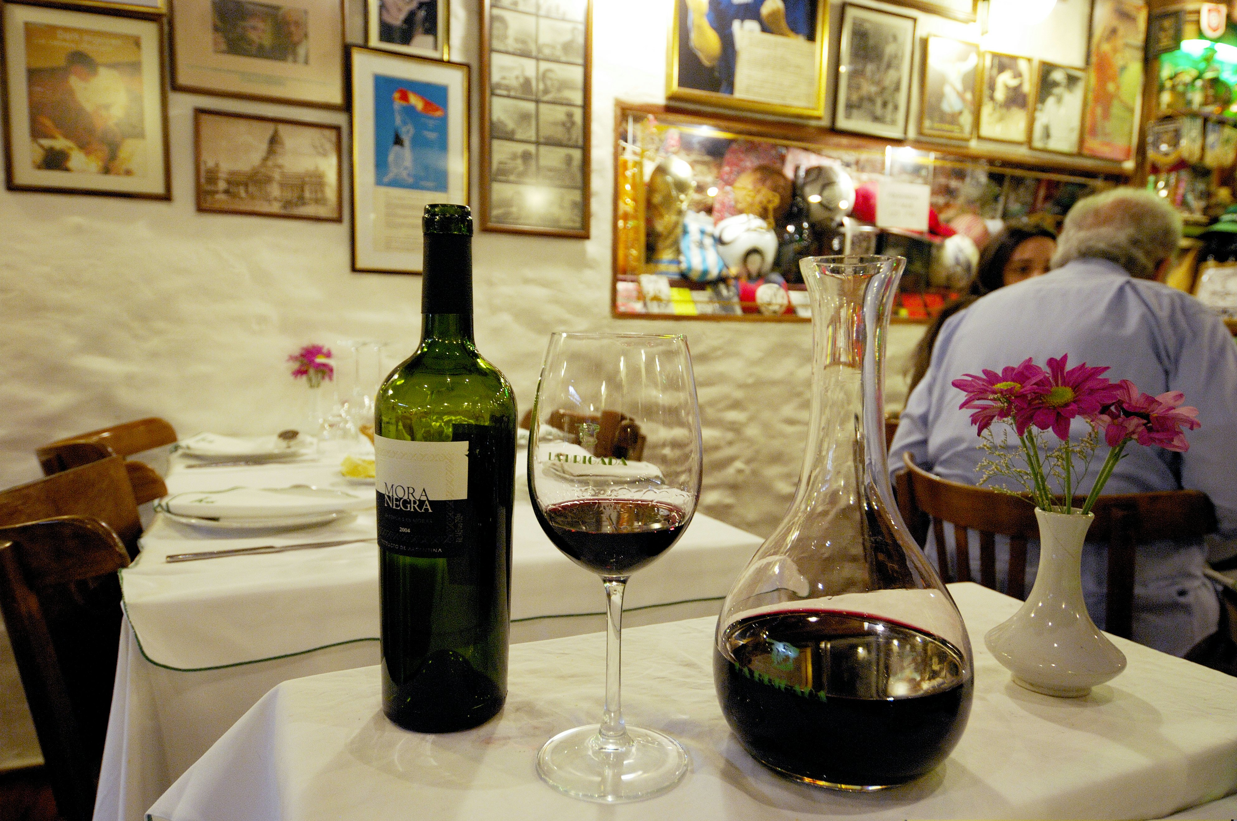 A bottle, glass and carafe all containing red wine are placed on a table at a restaurant. Colorful photographs and framed art can be seen on the wall in the background.