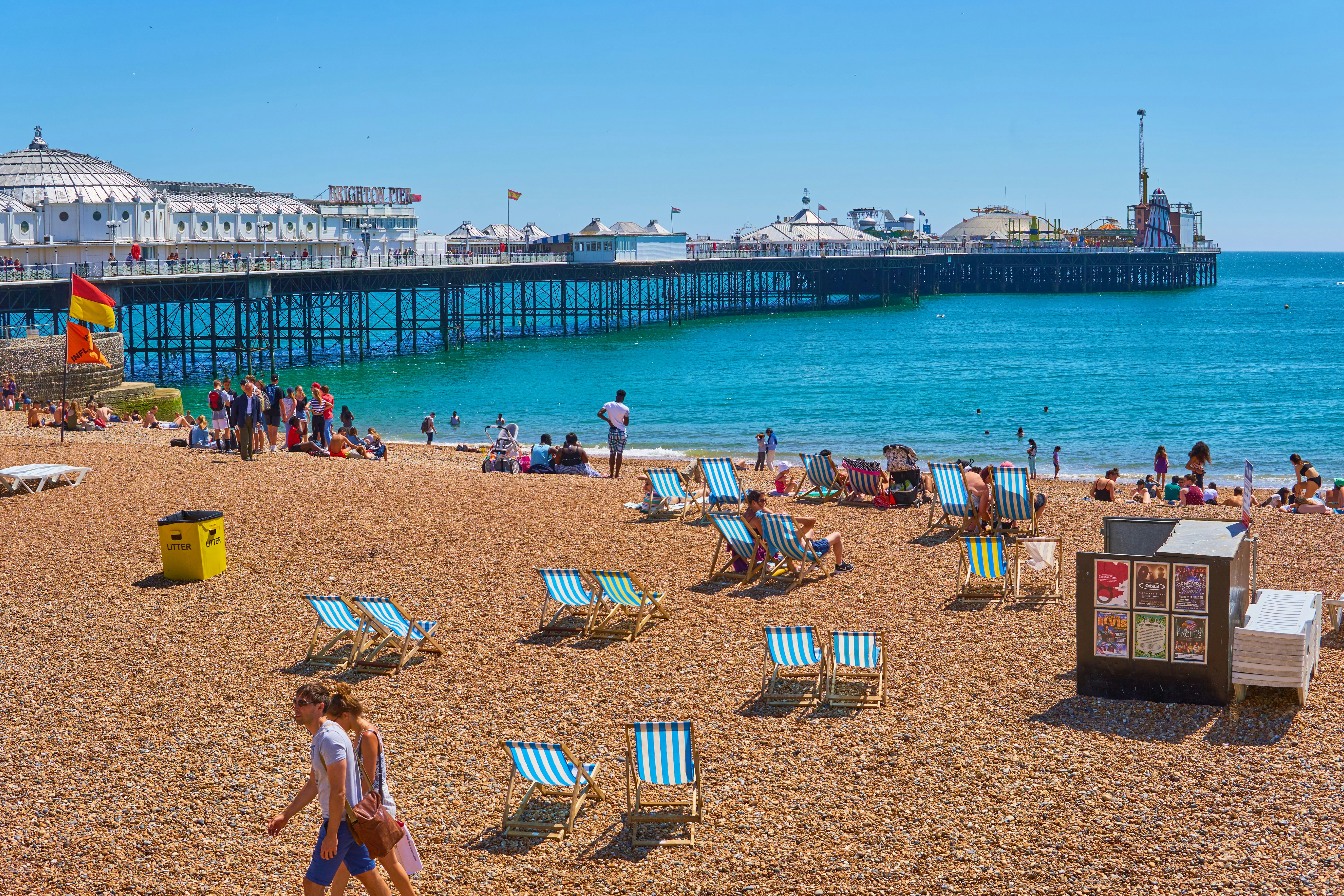 People lounge in deckchairs on pebbly beach near a large pier with amusements on a sunny day