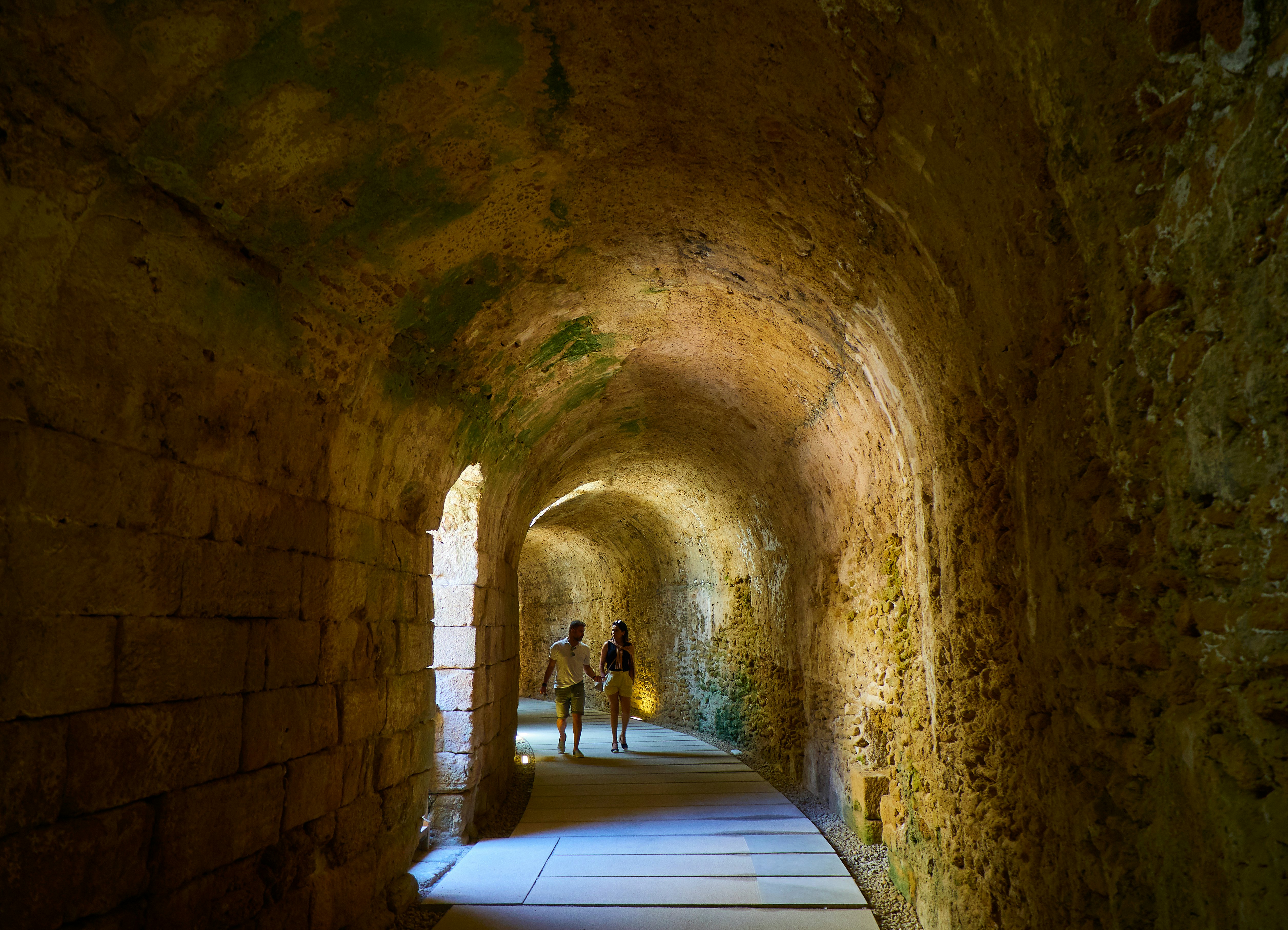 Two people walk down a curving corridor underground within an ancient structure.