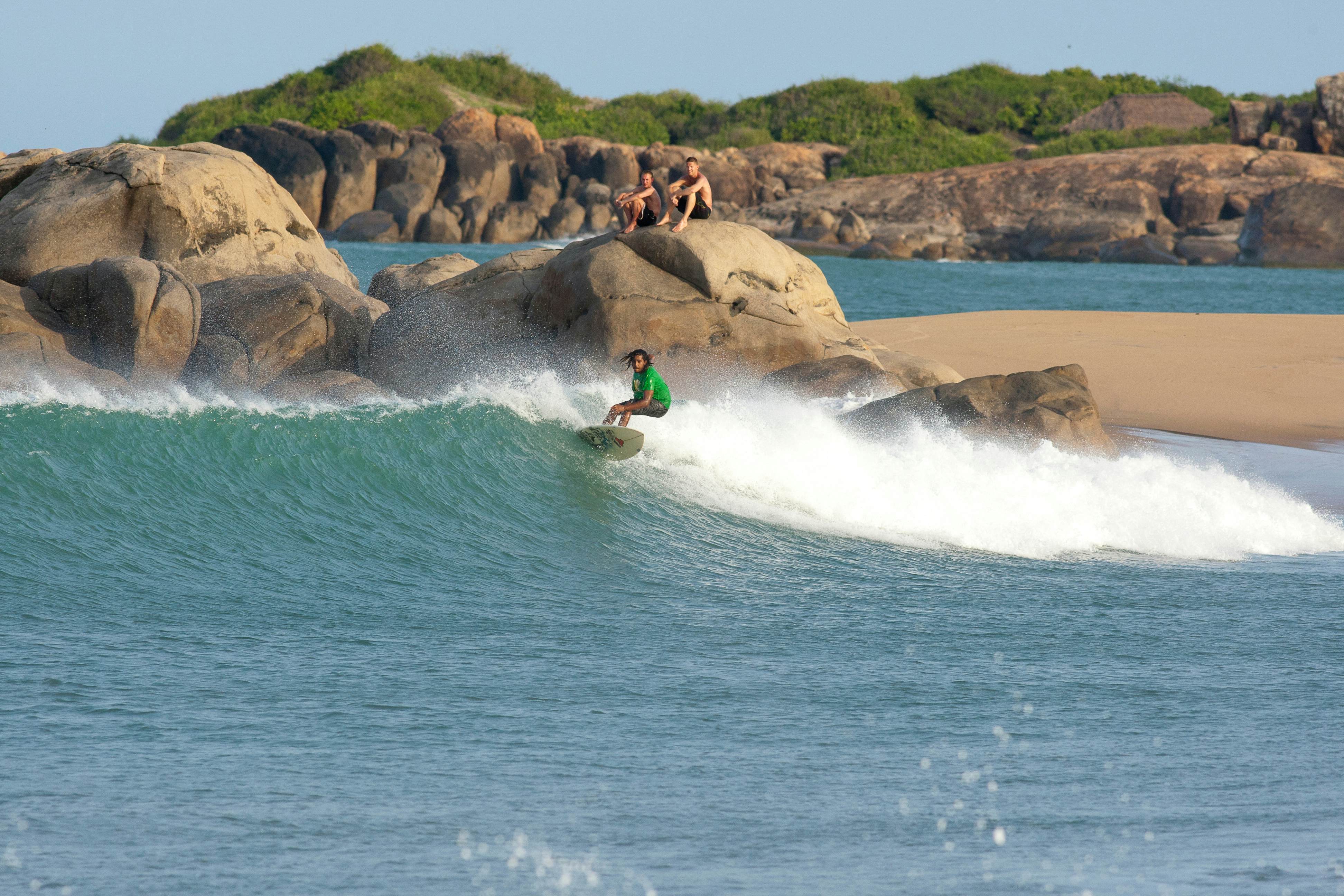 A local surfer rides a wave at a cove. People sit and watch on large boulders behind the surf break.