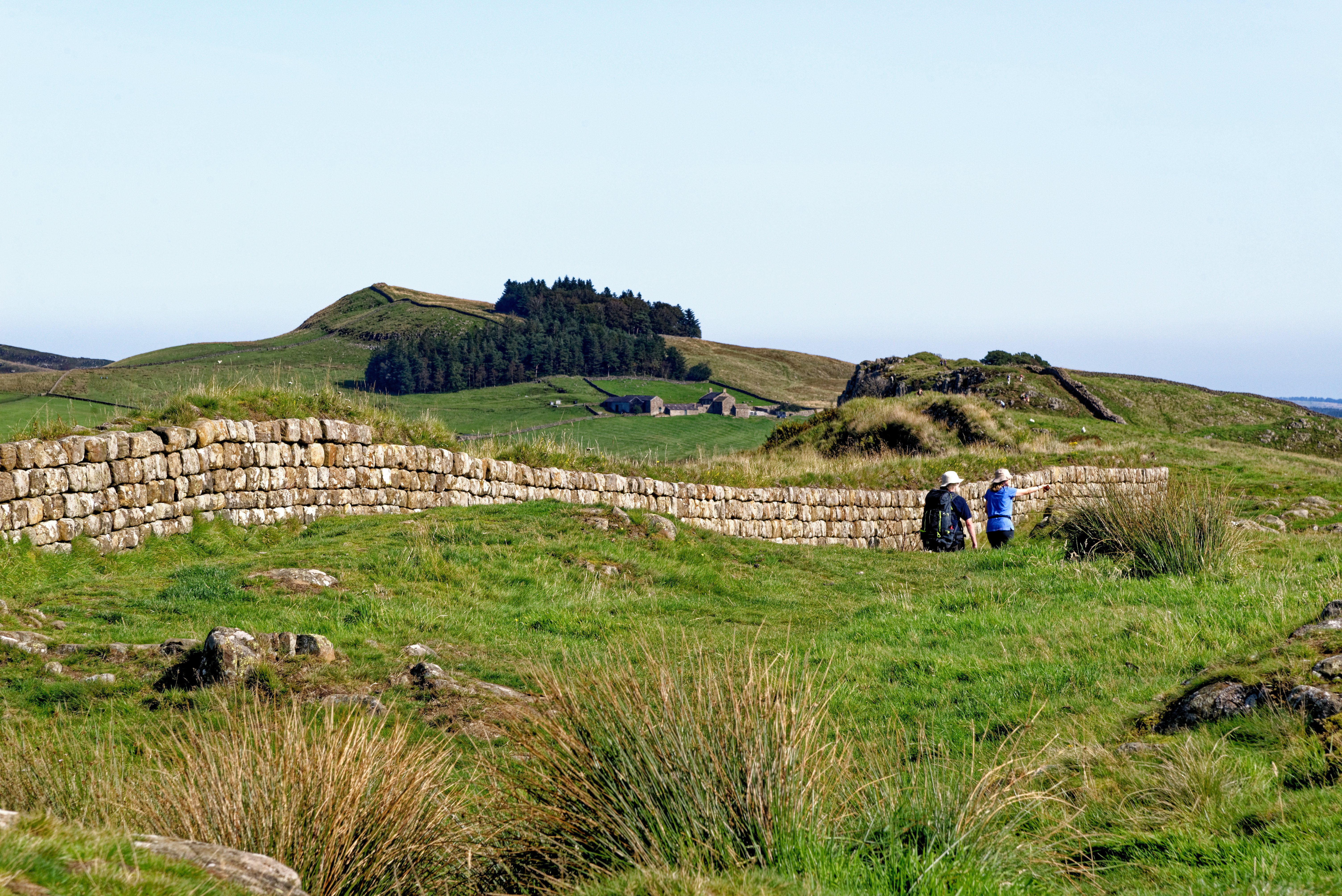 Two hikers walk through fields beside an ancient all that continues over hills and far into the distance