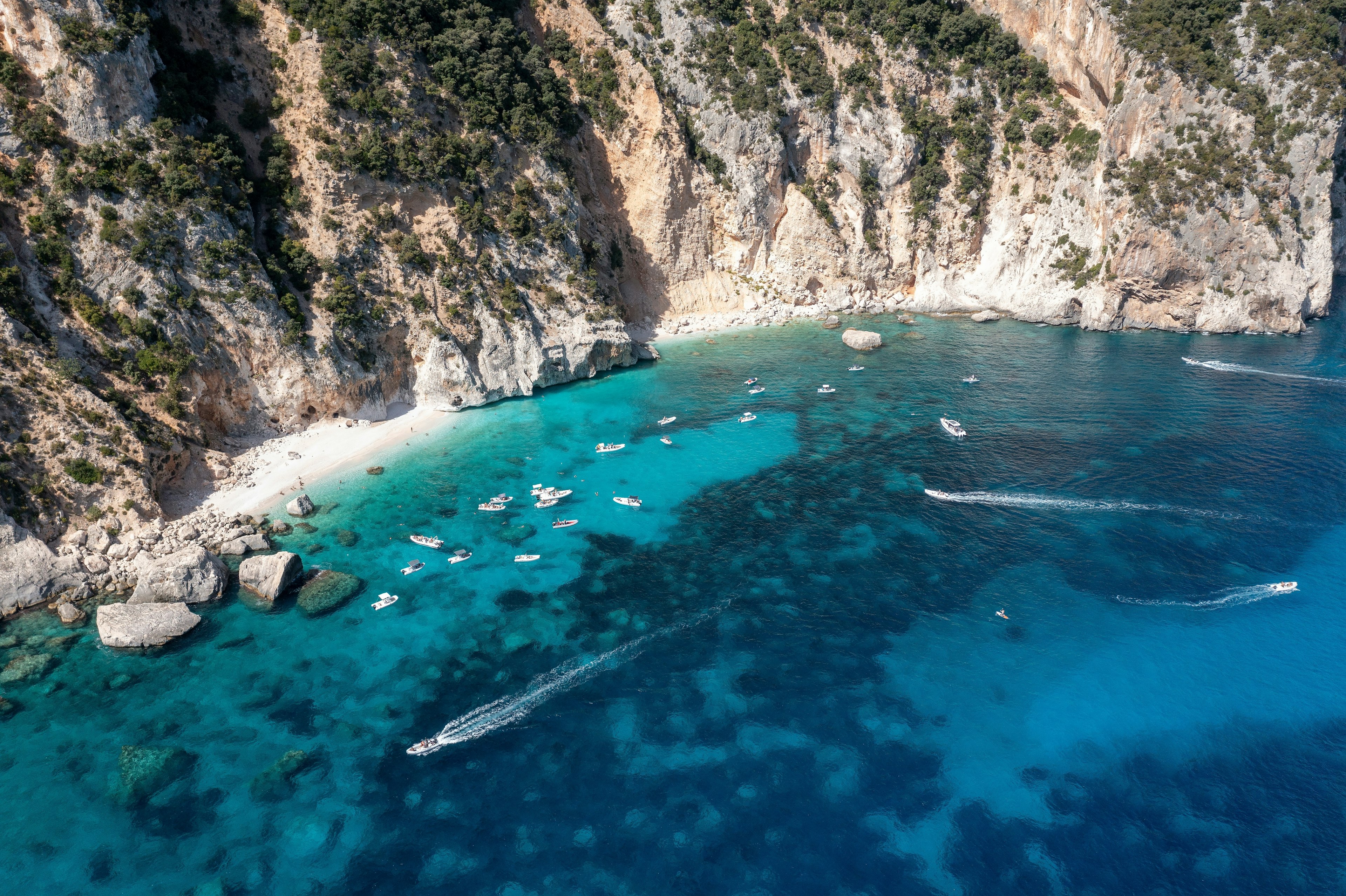 An aerial view of boats moored at cove with small beaches against a steep, rugged cliff.