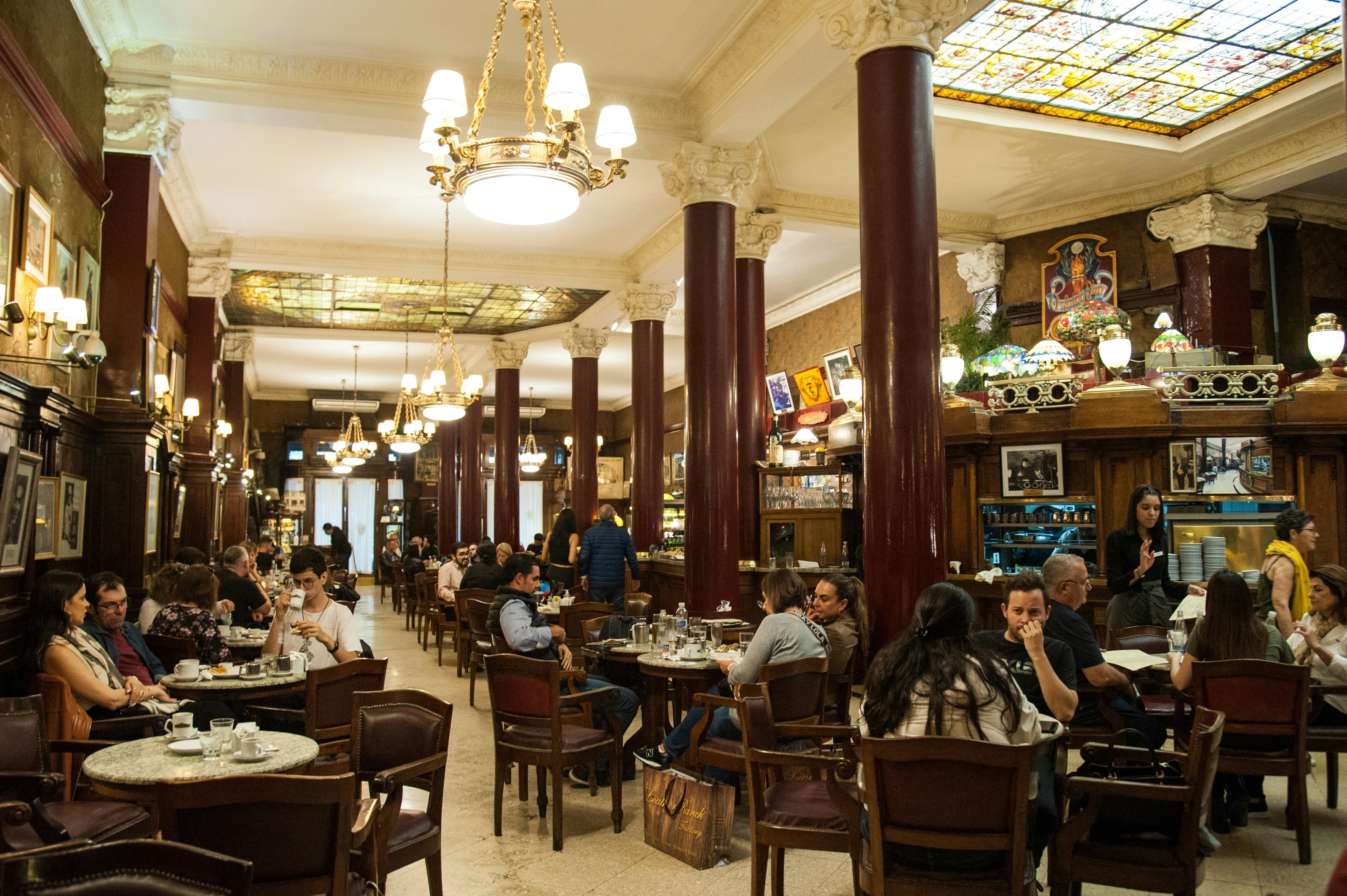 Patrons sit at tables in the historic dining room of a restaurant, with dark-wood chairs, chandeliers and stained-glass panels in the ceiling.