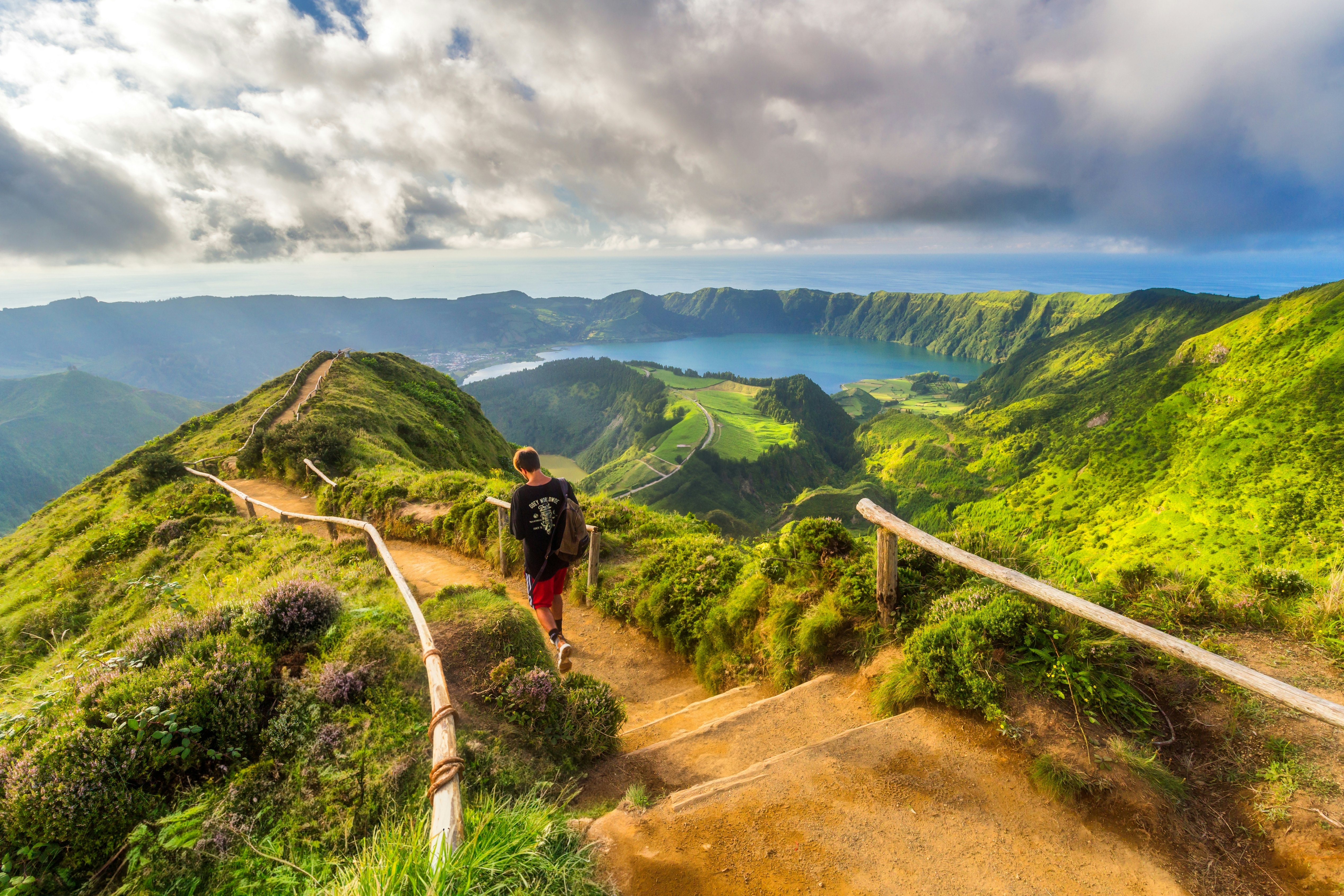 A hiker with a backpack walks down a dirt path following the ridge of the hill overlooking a volcanic lagoon. The surrounding hillsides and cliffs are green and lush.