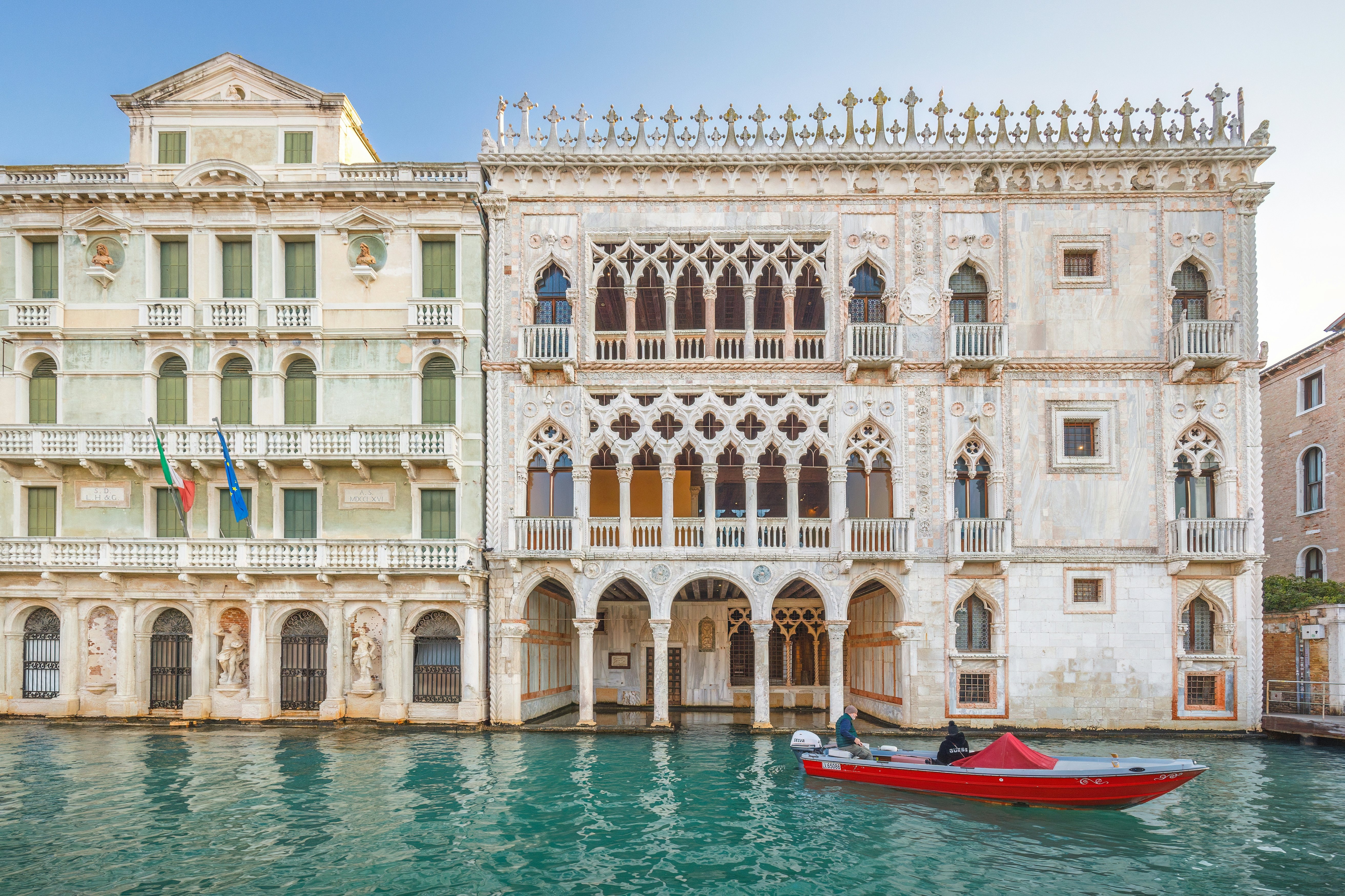 Ornate facade of a palace with balconies that appears to sit on the water of the canal city. A red boat is passing by