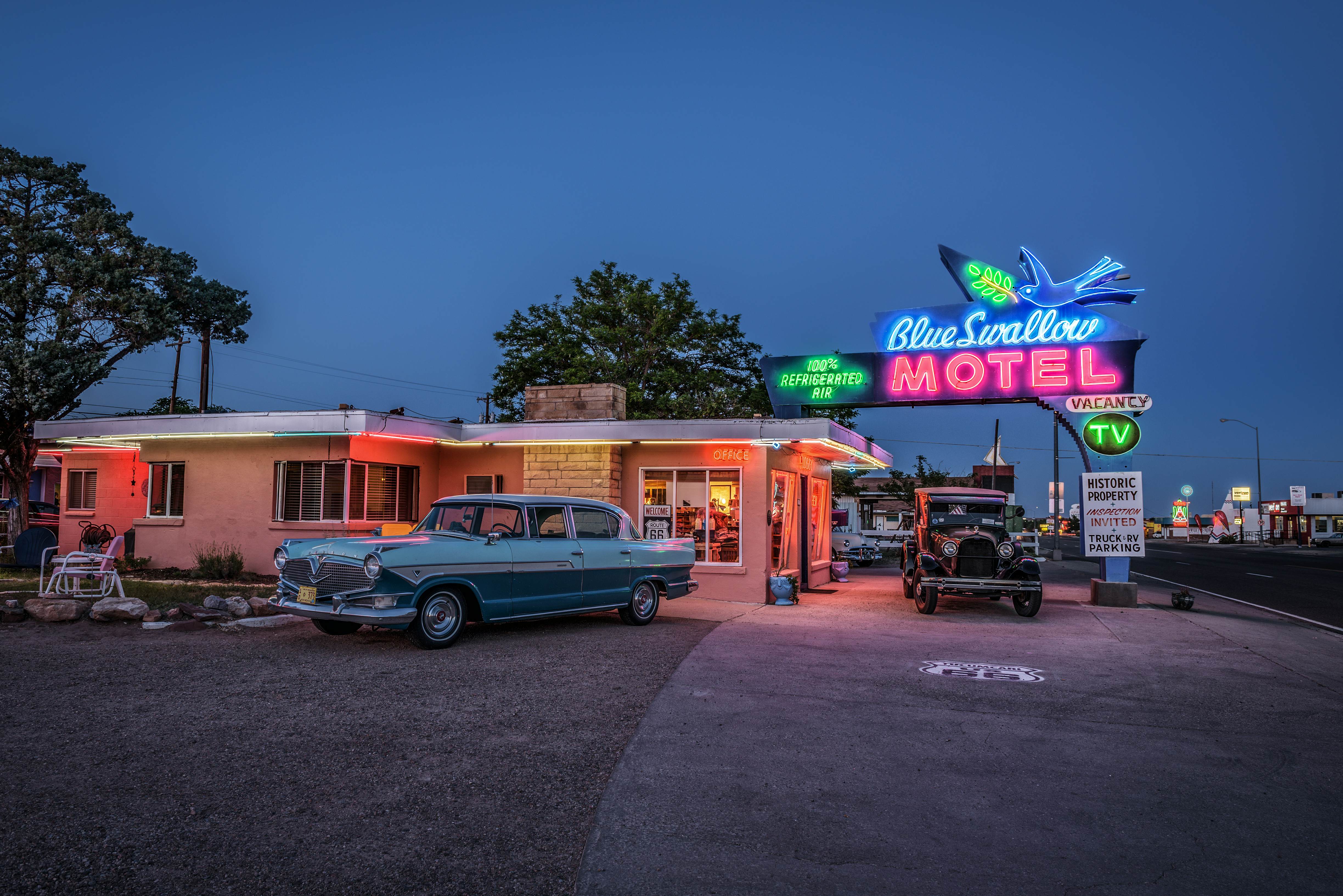 Two vintage cars are parked in the driveway of a motel illuminated in the evening by neon lights.