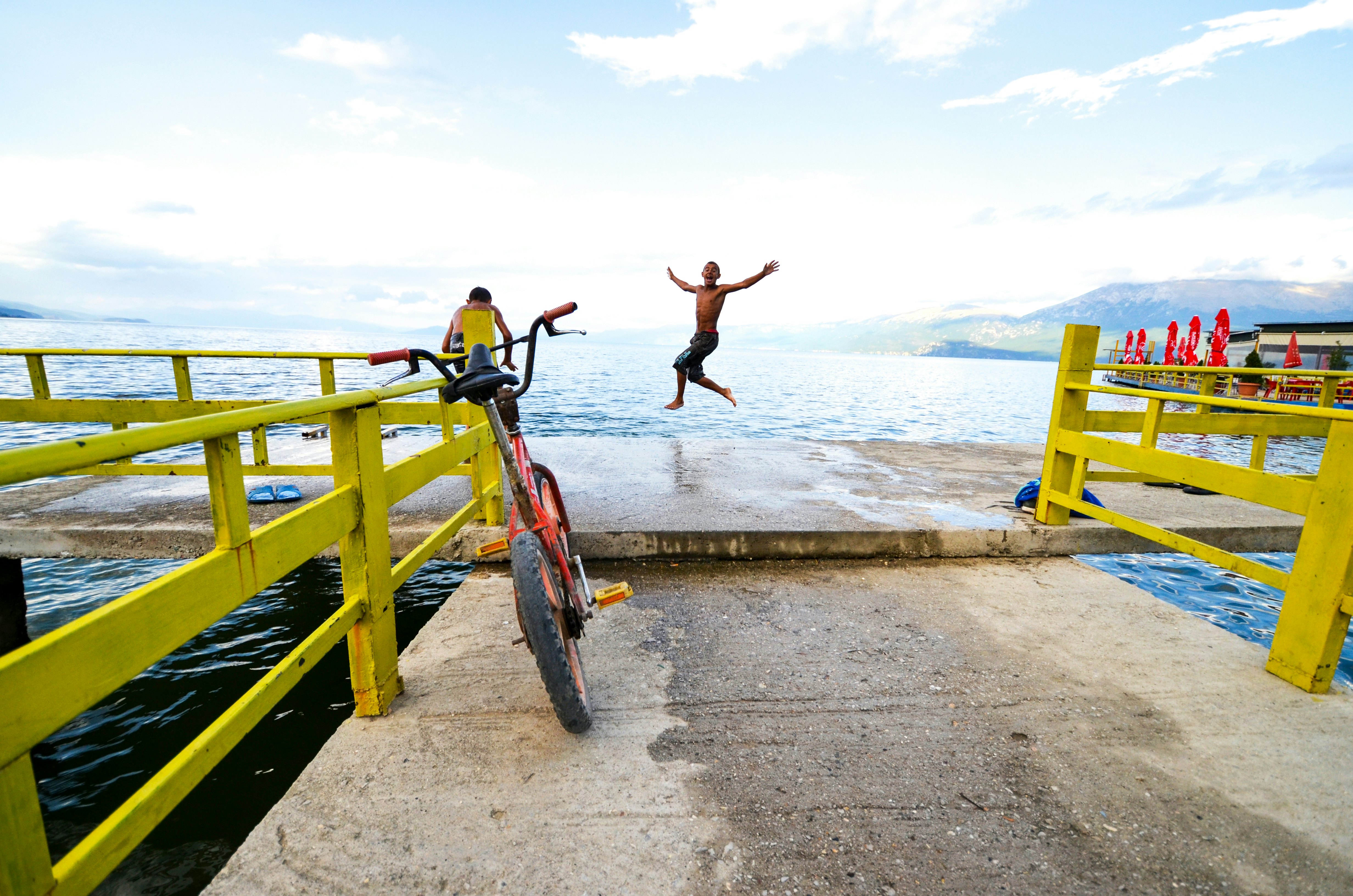 A teenager jumps into the water of a lake in a mountain setting. In the foreground, a bicycle is propped against a yellow fence.