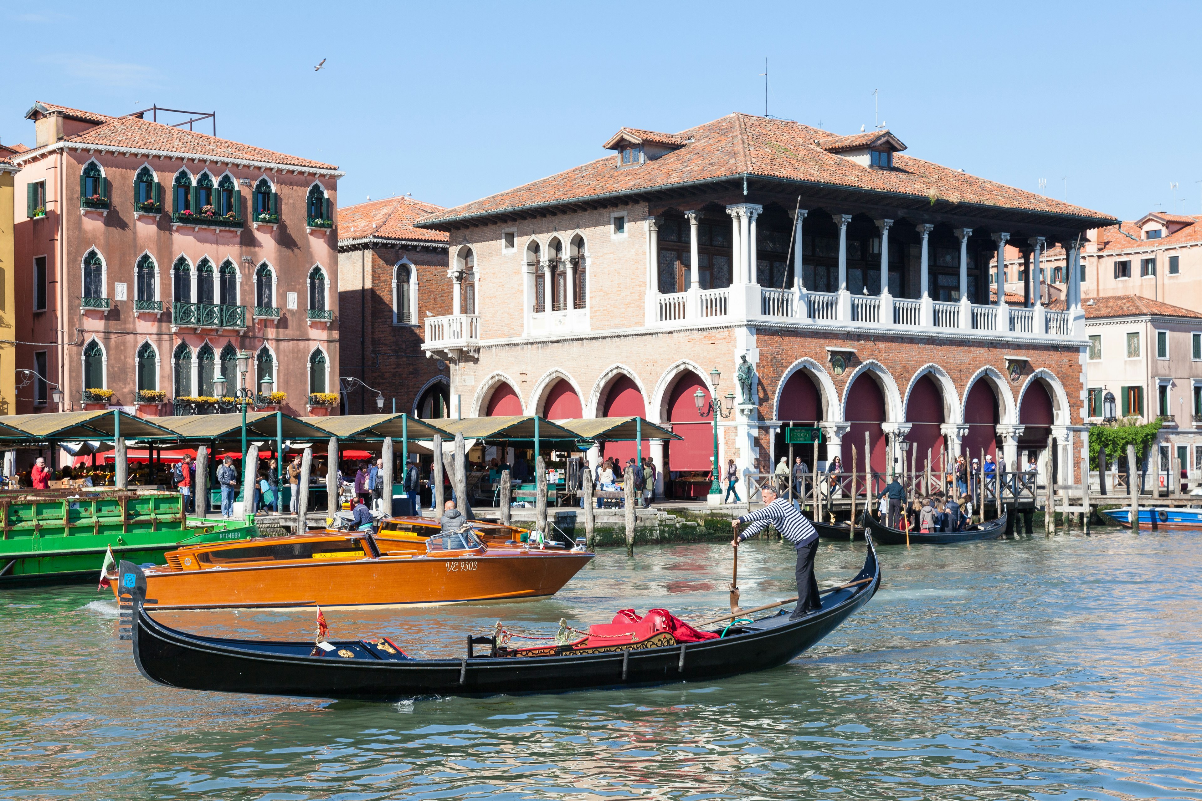 Gondolier and gondola on the Grand Canal in front of the historic Rialto Market with shoppers buying  ish and vegetables at the stalls and a water taxi passing