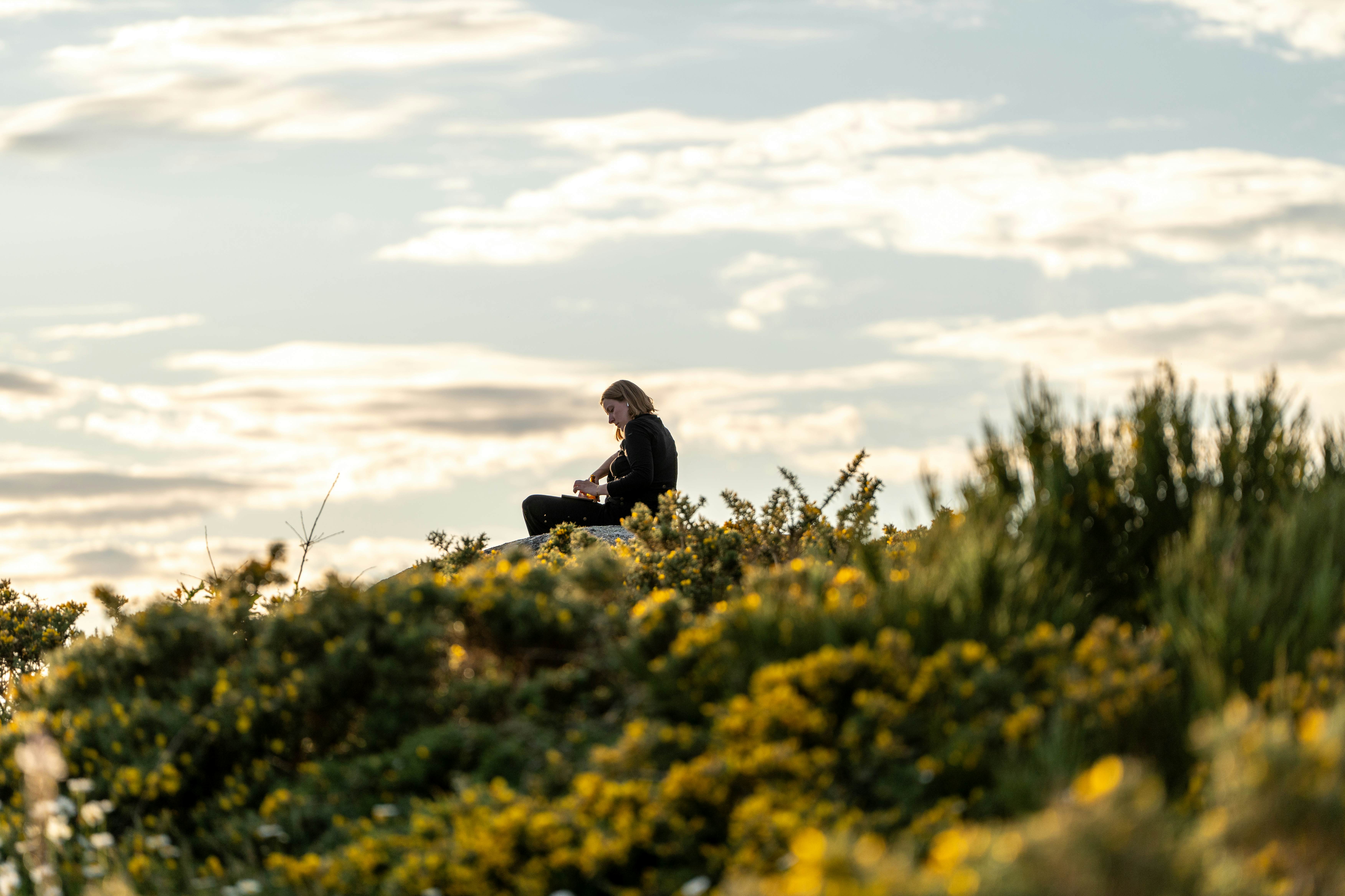 Woman sits in gorse at the edge of a cliff in Finisterre to watch the sunset.