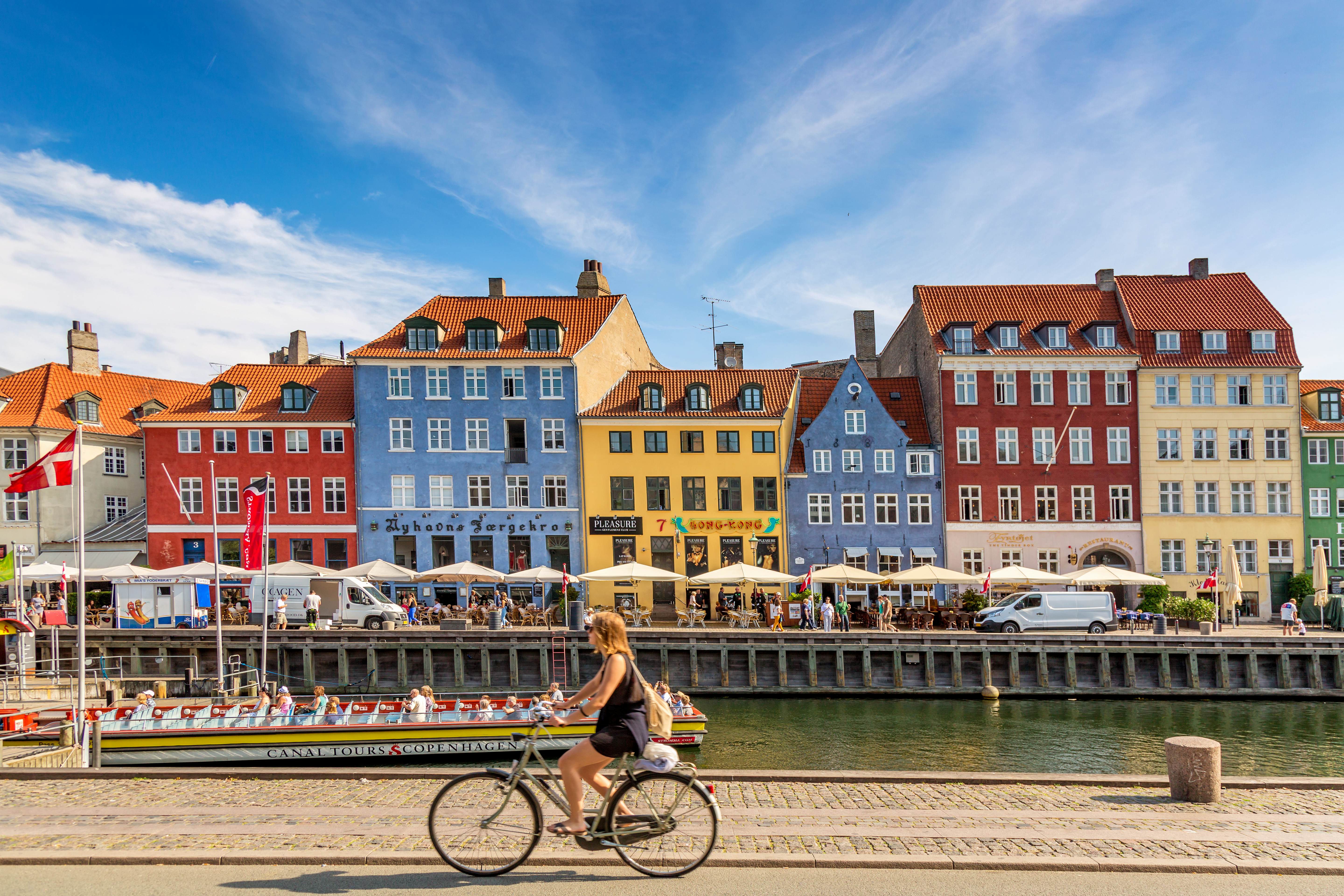 A woman rides a bike along a harbor lined by buildings painted in shades of red, blue and yellow