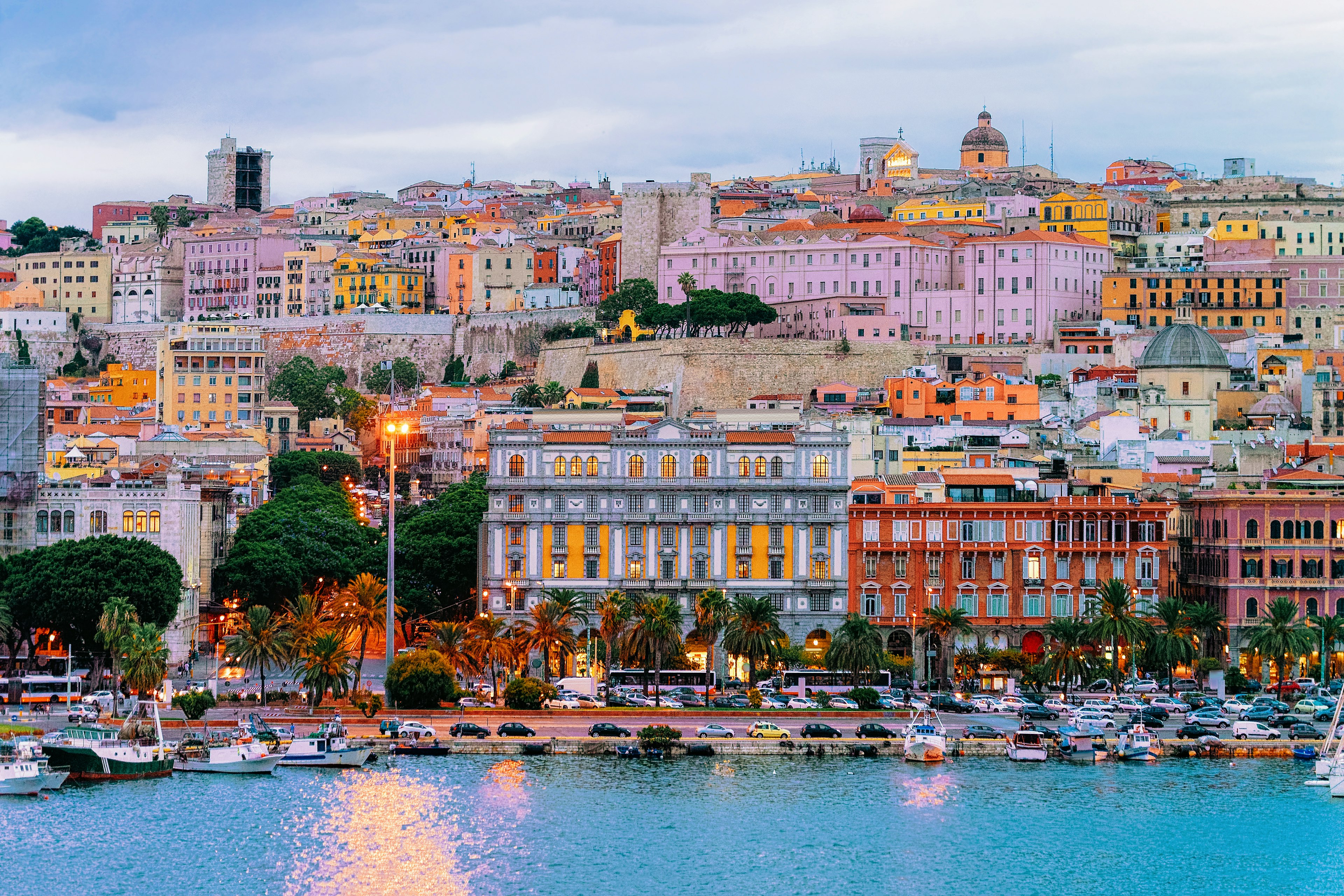 A view from the water of the colorful facades of buildings rising on a hill in a city. Boats are visible in the marina, and a road leads along the waterfront.