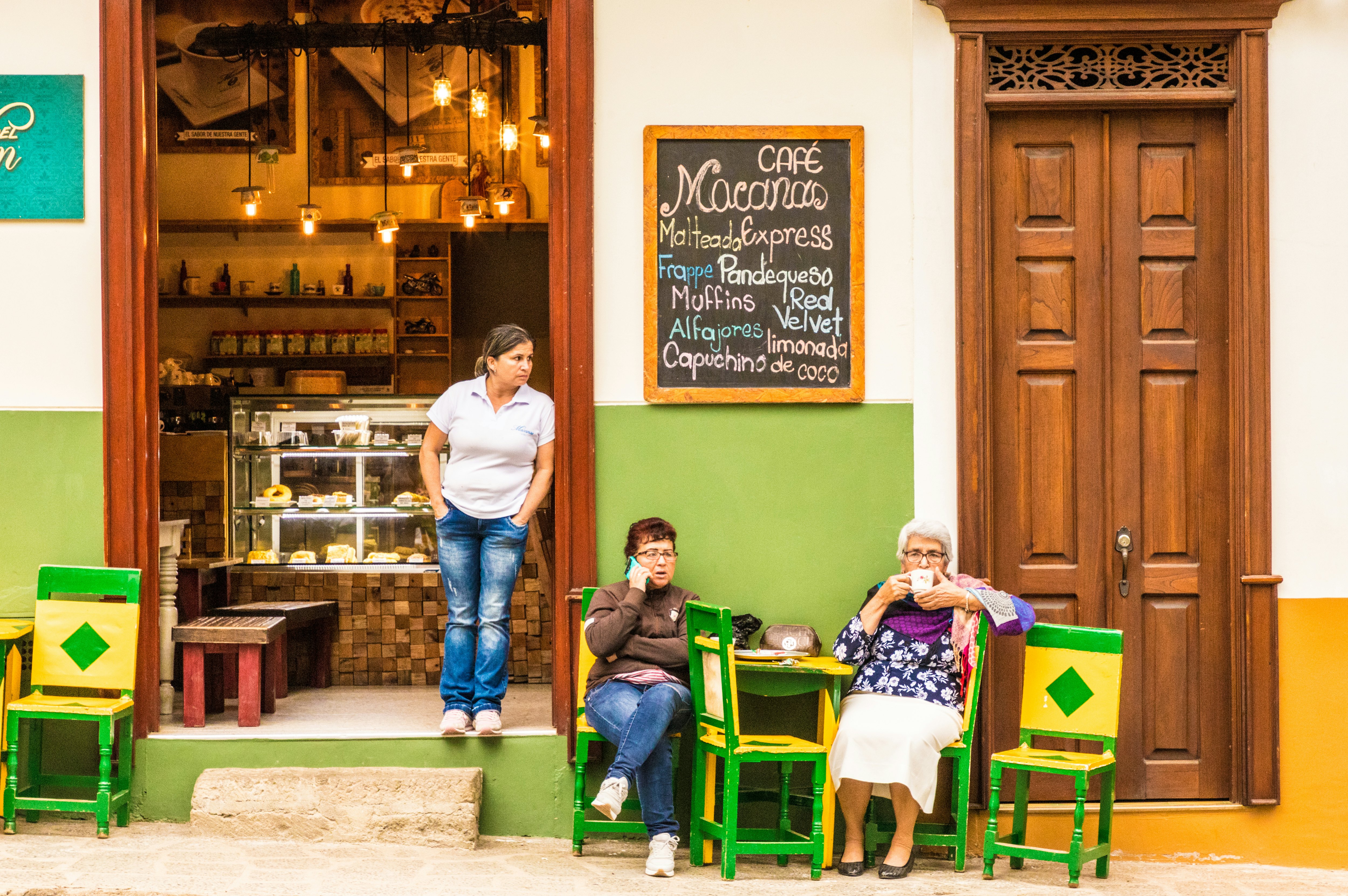 Three women are pictured at a cafe on the street level of a historic buildings. Two sit at a table. The tables and chairs are painted green and yellow.