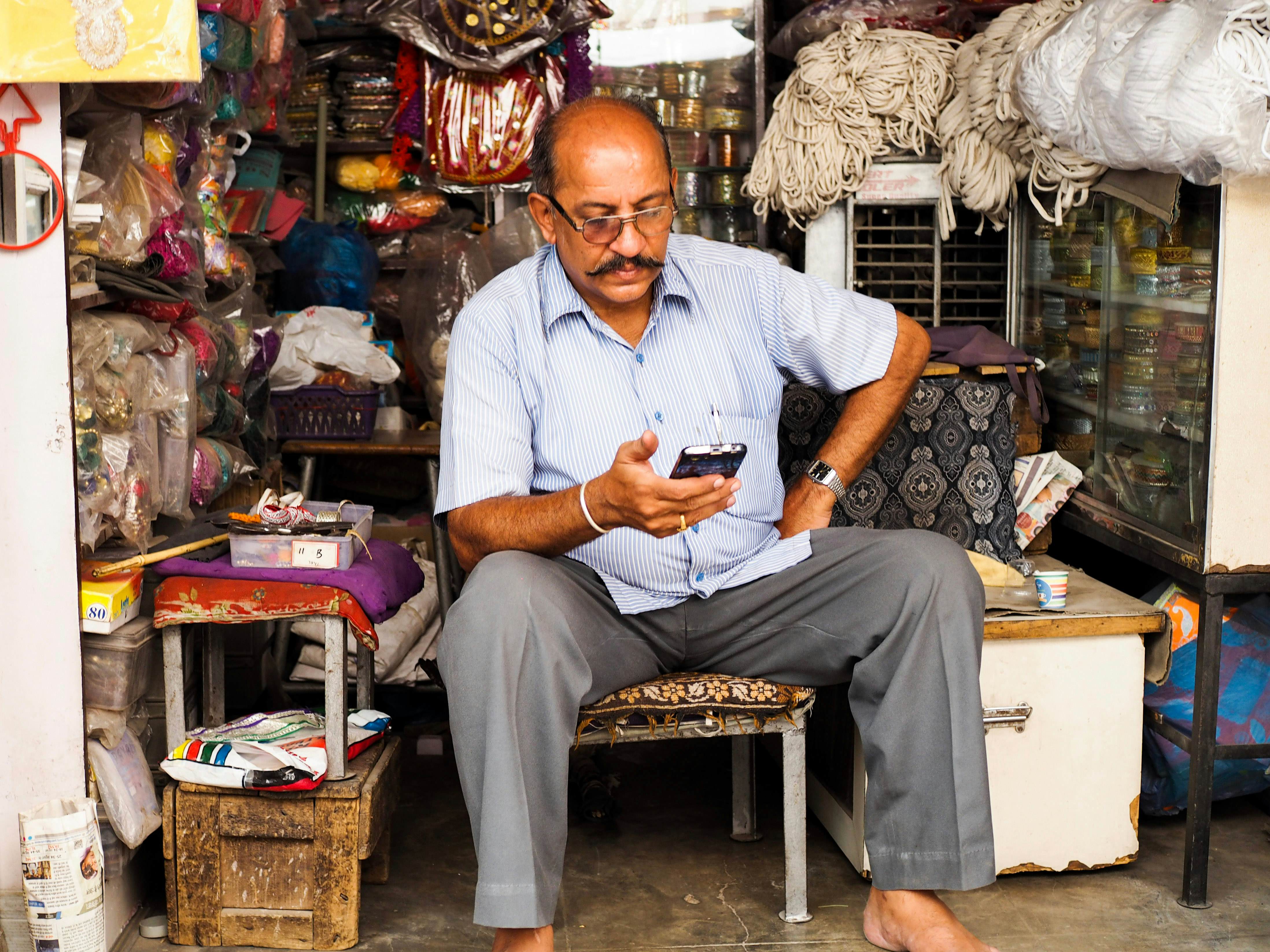 A man sits at his small shop and looks at his phone.