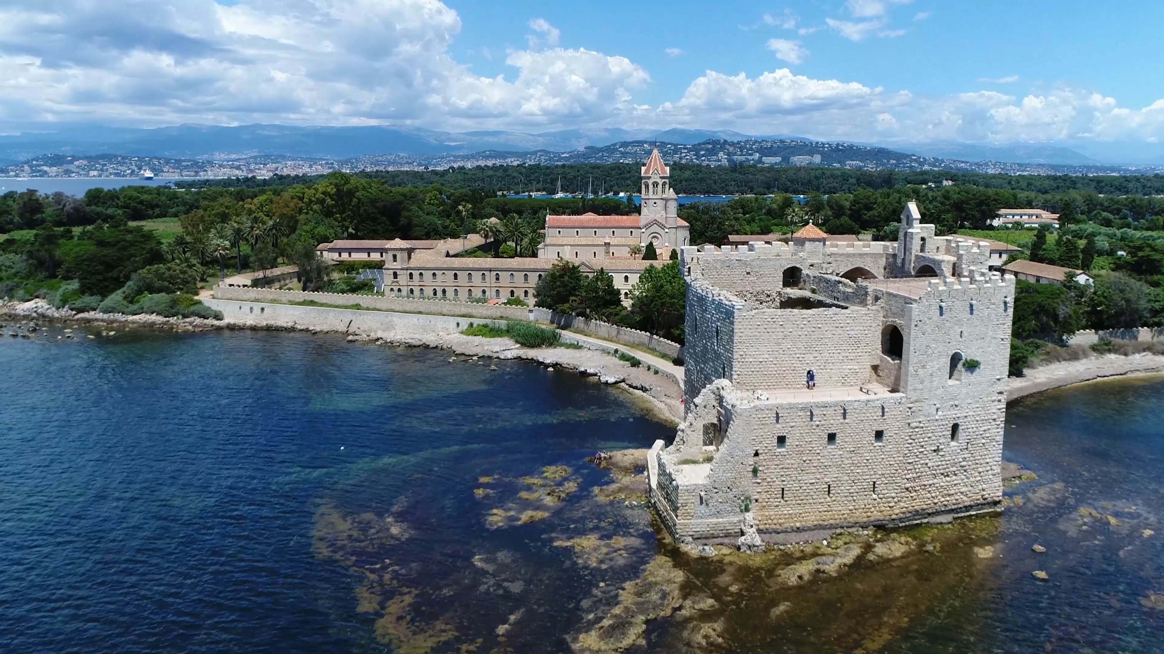 An aerial photo of a historic fortress and monastery complex on an island. The mainland, with buidlings and boats offshore, is visible in the far distance.