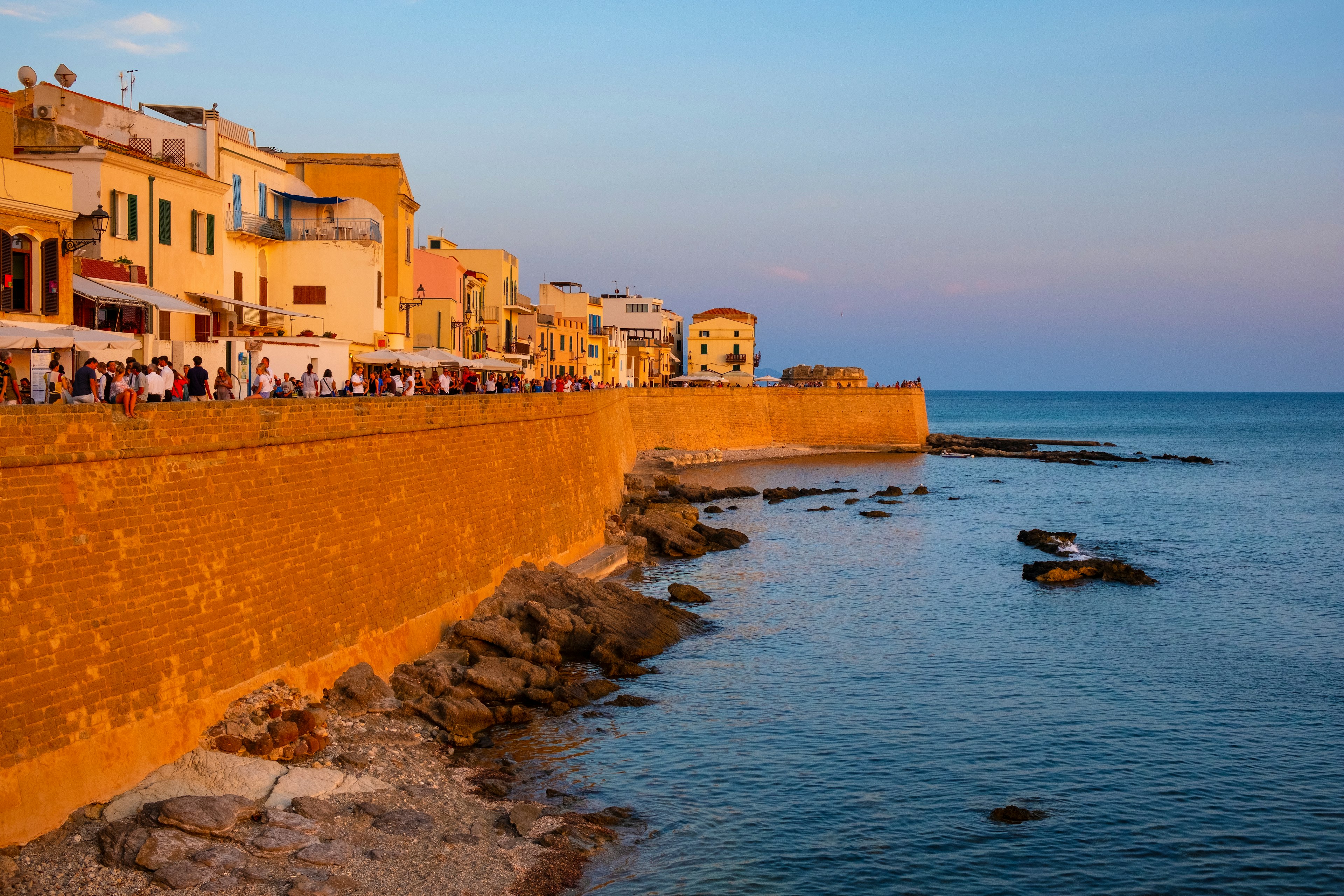 People line the sea wall of a historic city to watch the sunset. Rocks are visible in the water that abuts the wall.