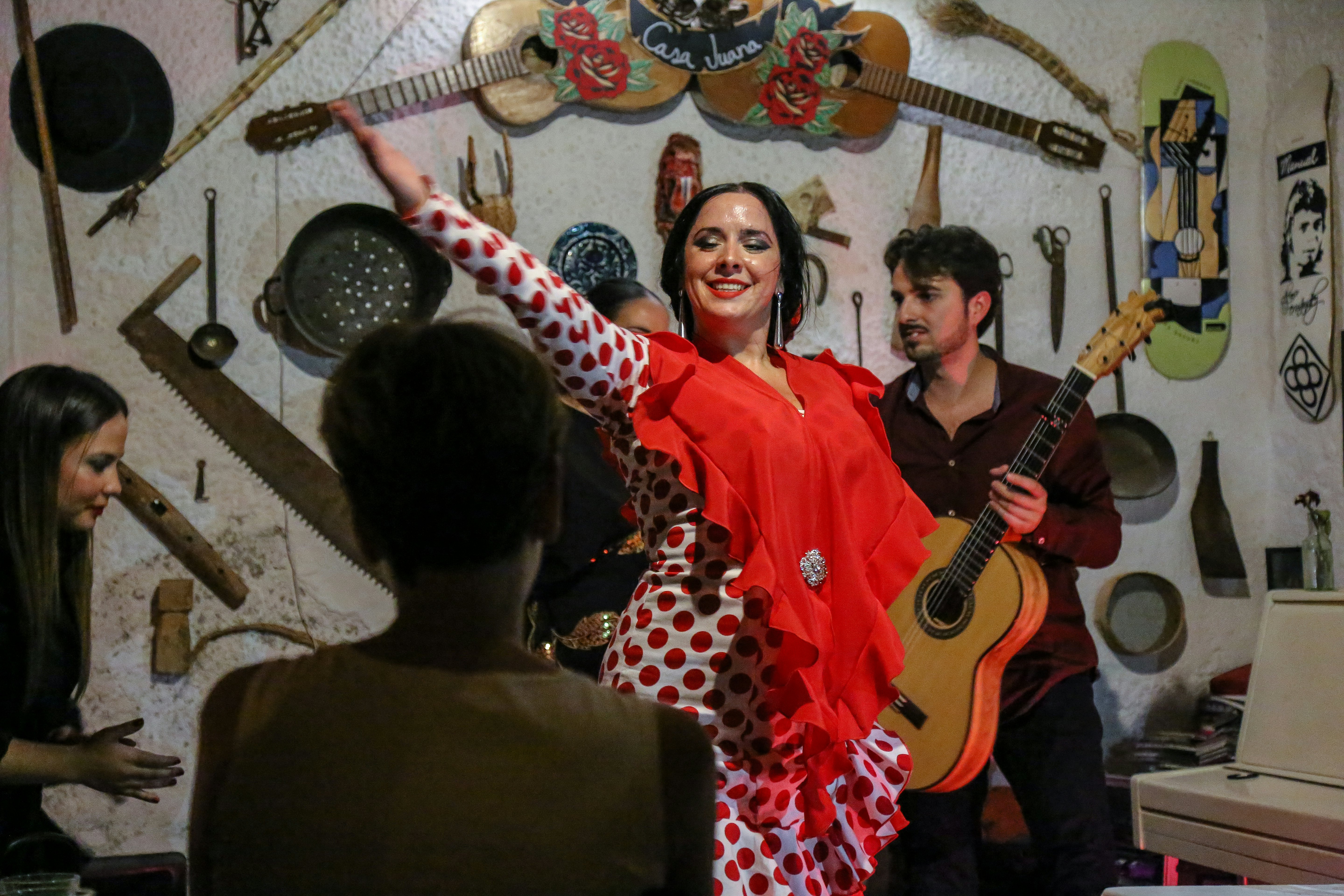A flamenco dancer in a white dress with red polka dots wears a red shawl; musicians are playing behind her in front of a white wall with instruments and tools hanging on it.