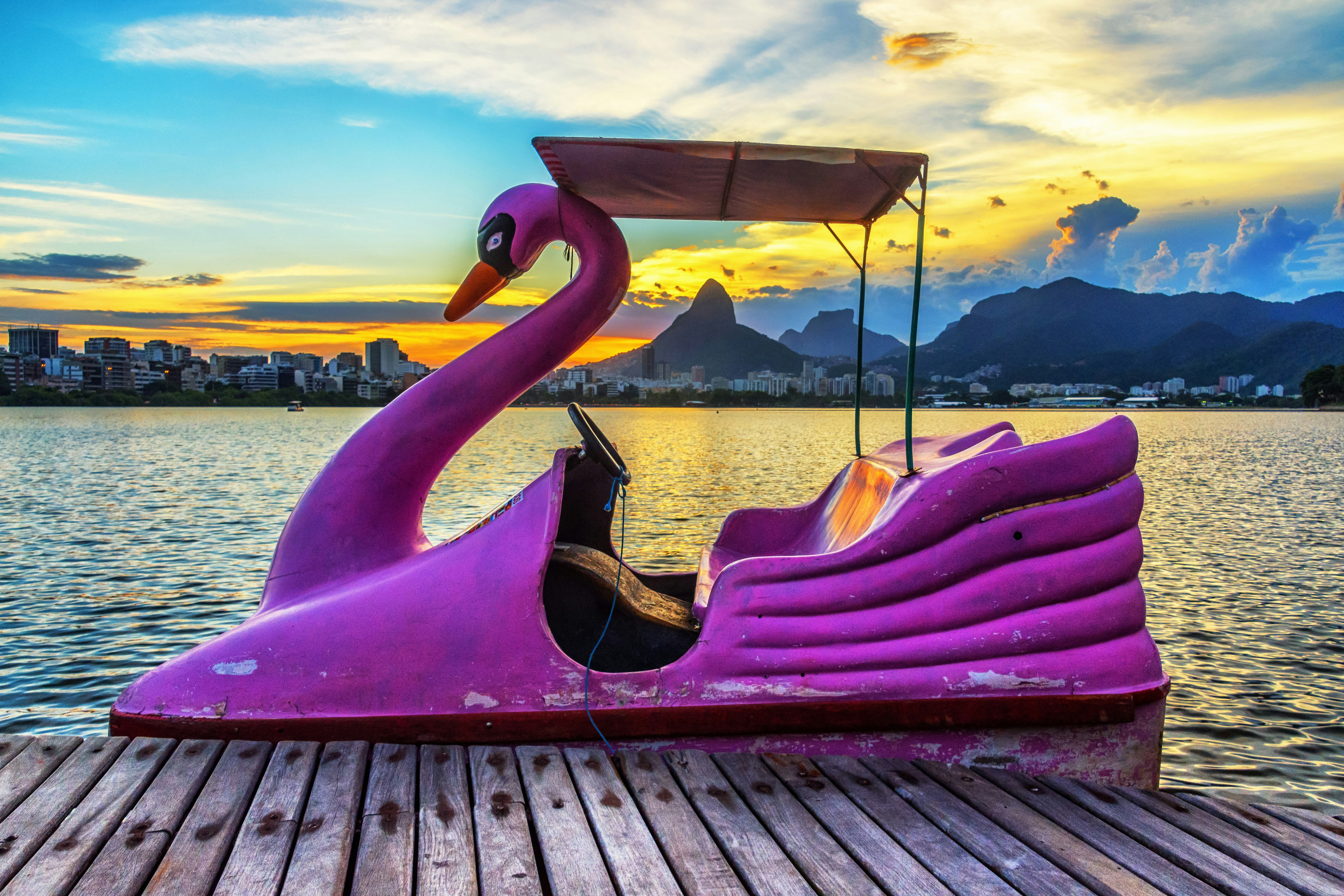 A flamingo-shaped pink pedalboat sits at a dock on a lake at sunset. Mountains and a city skyline are visible across the lake.