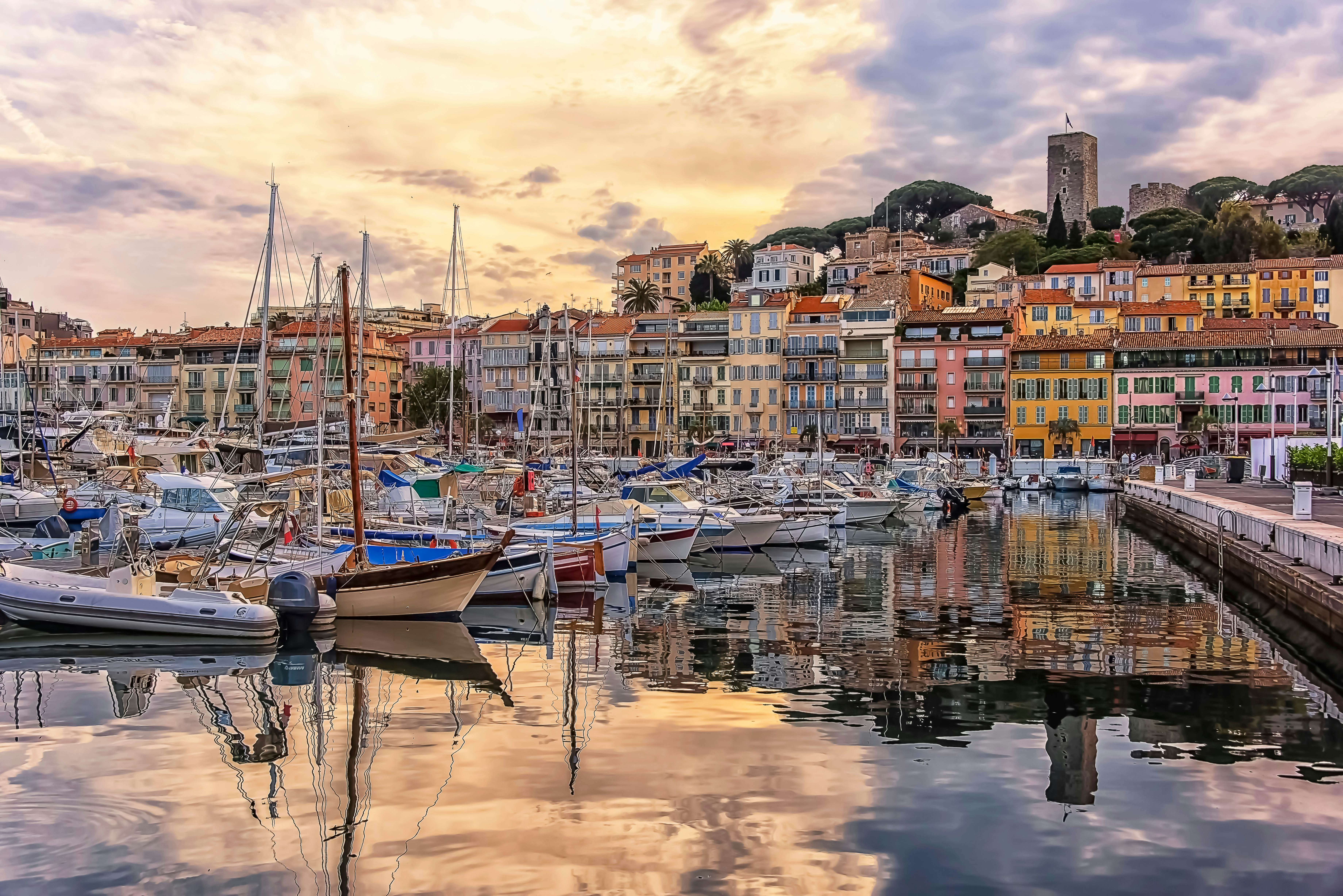 Boats are moored in a port at sunset. Their outlines are reflected in the water. A line of colorful buildings lines the shore, and a castle sits on a hill that rises behind them.