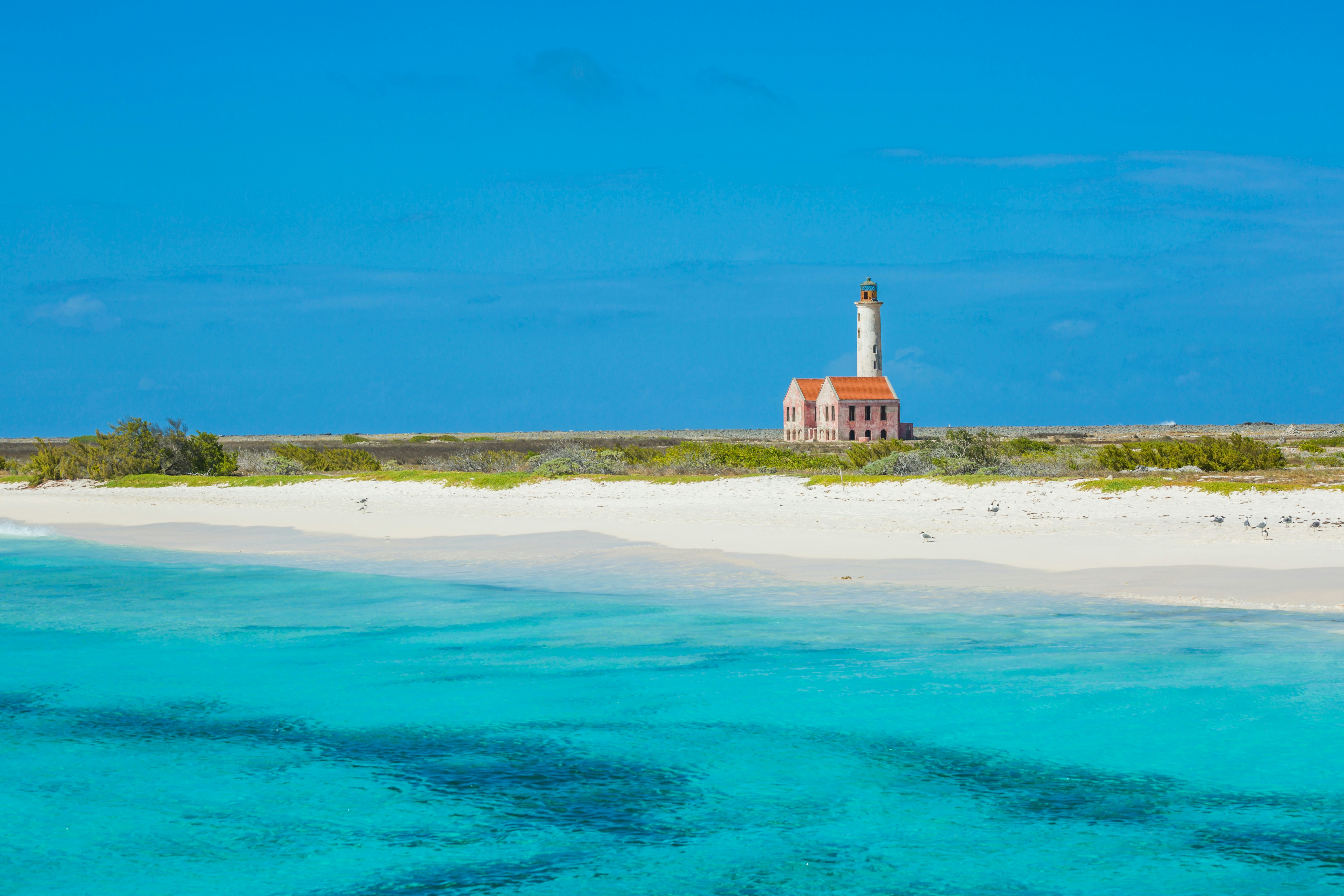 A lighthouse stands alone on a very flat island, with blue ocean and sparkling white sand.