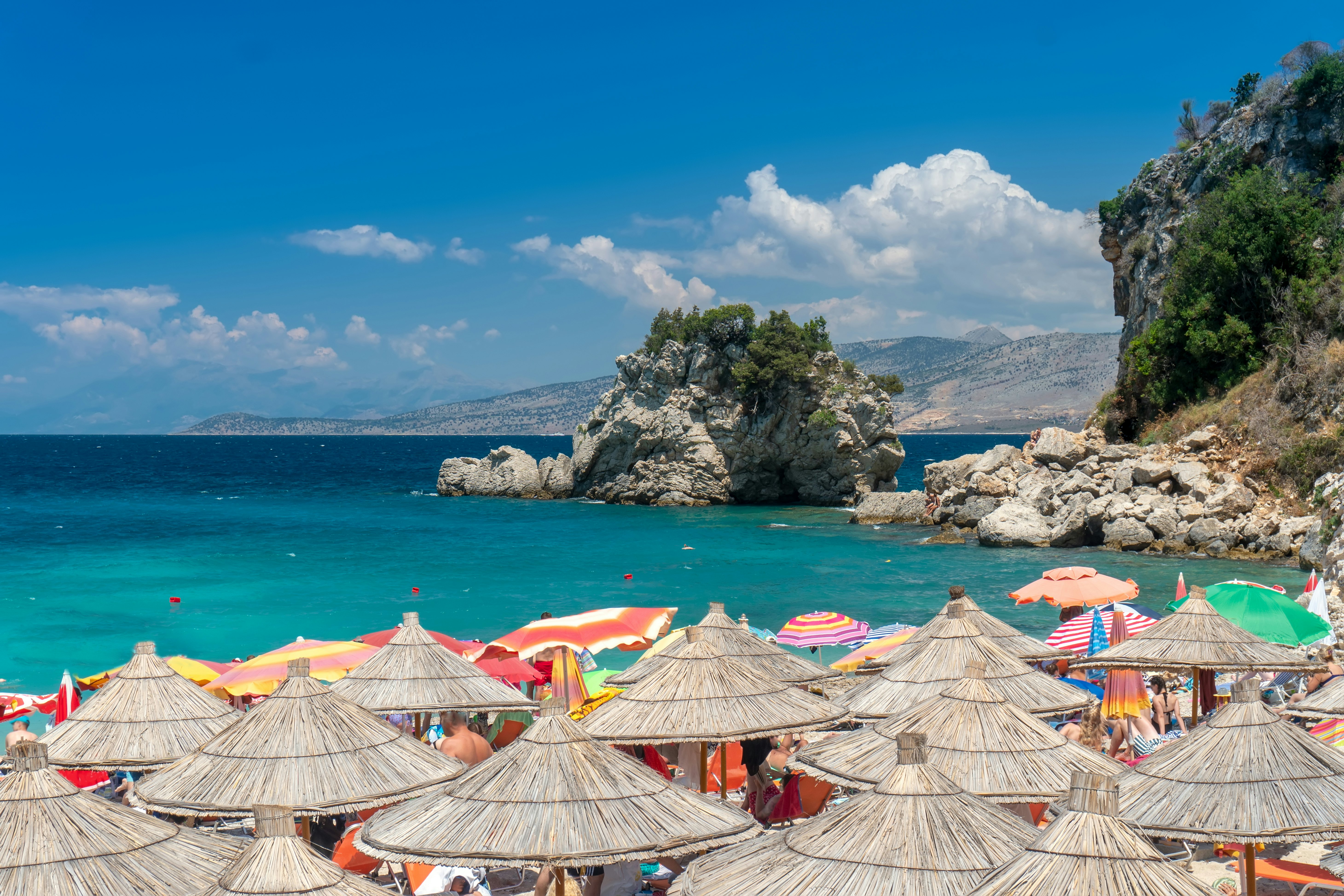 The tops of numerous straw sun umbrellas are pictured on beach. Turquoise-colored water and rocky islands are seen in the distance.