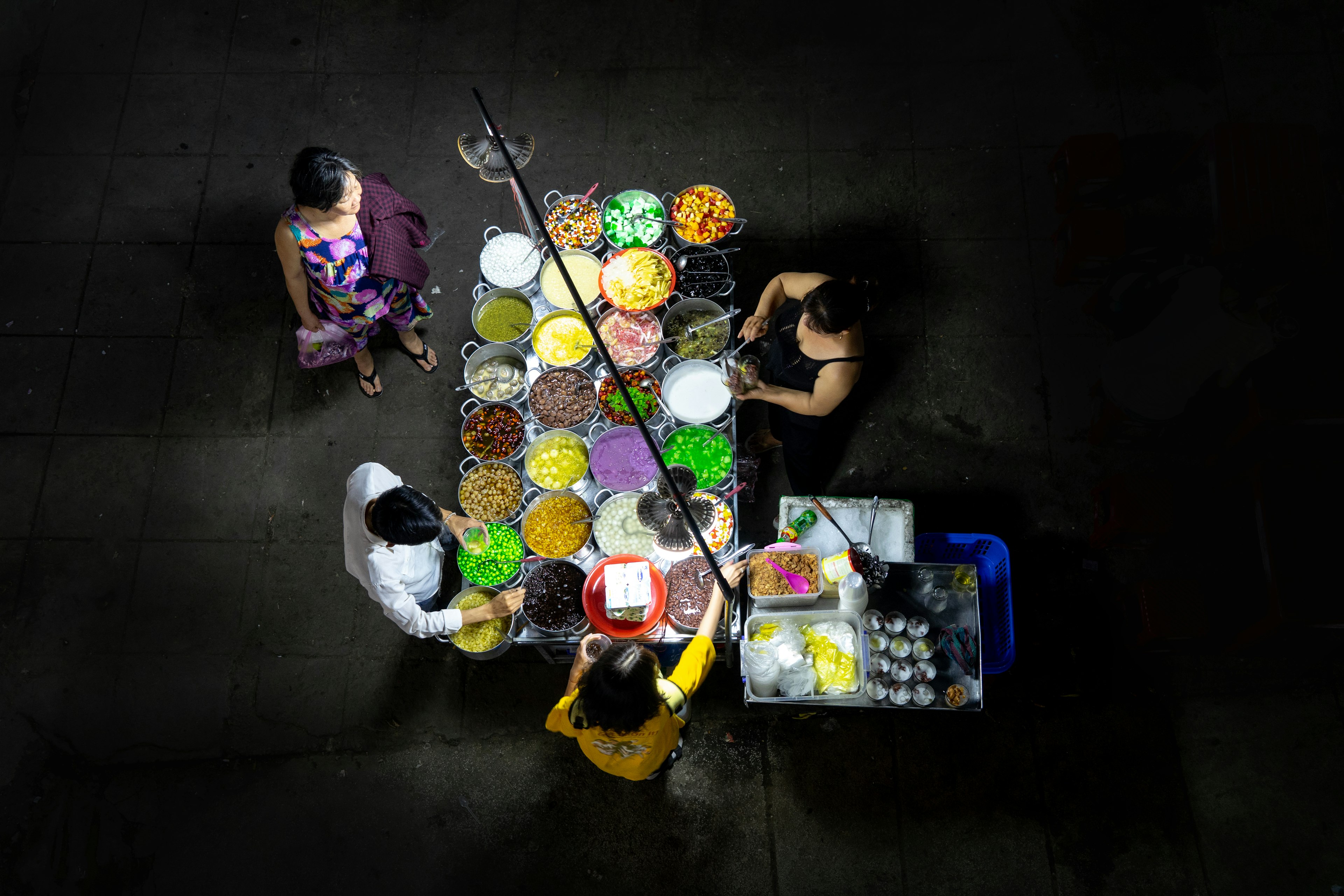 An aerial view of a street vendor selling food at night, with colorful ingredients in containers brightly lit. Three customers are visible at the stand.