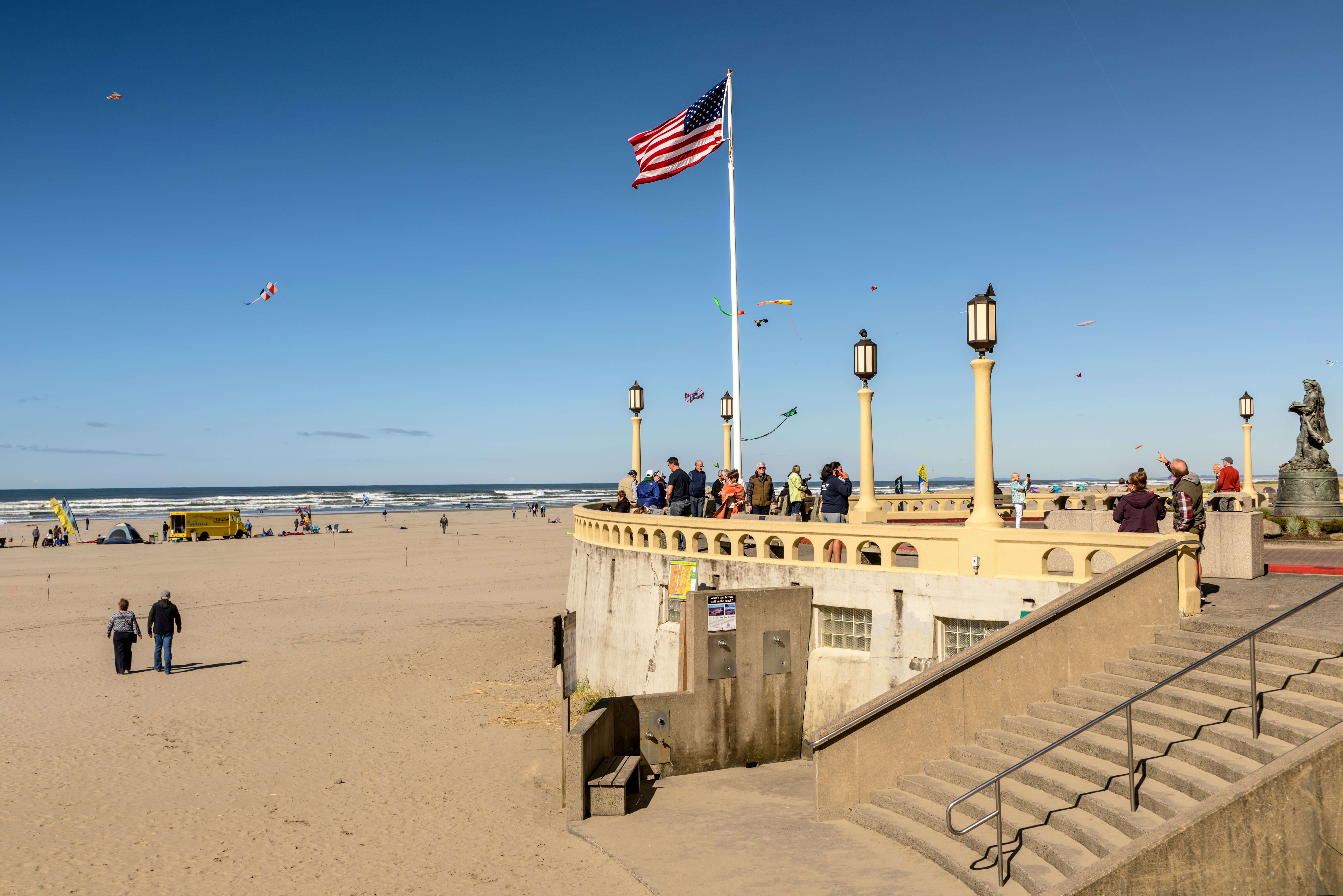 People watch a kite festival from the prom beside a sandy beach