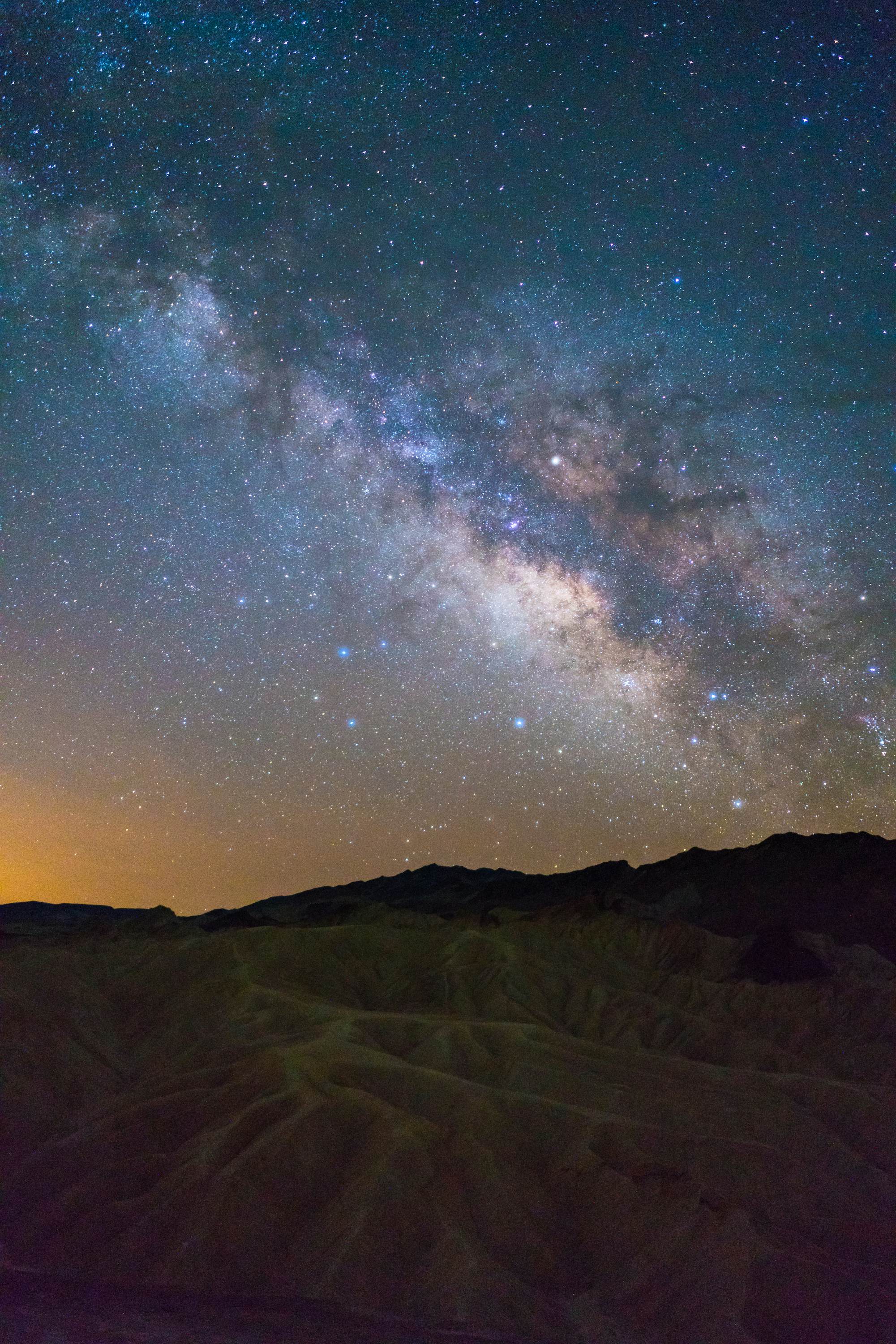 Milky way over a rippling rock formation with a distant orange glow in the sky