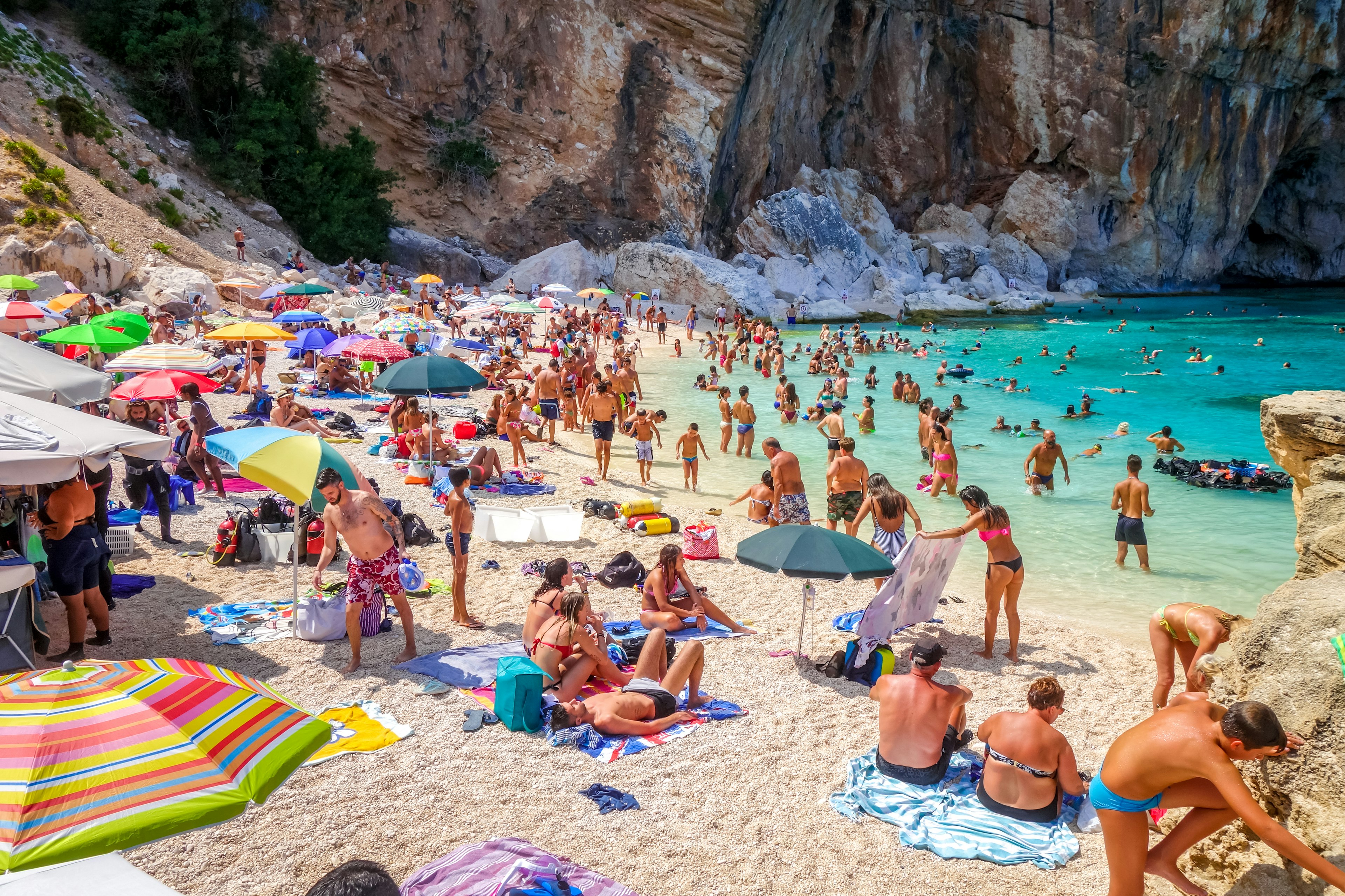 People in swimsuits crowd on a beach, many with sun umbrellas. Dozens of people swim in the shallow blue waters, and a tall cliff is visible in the distance.