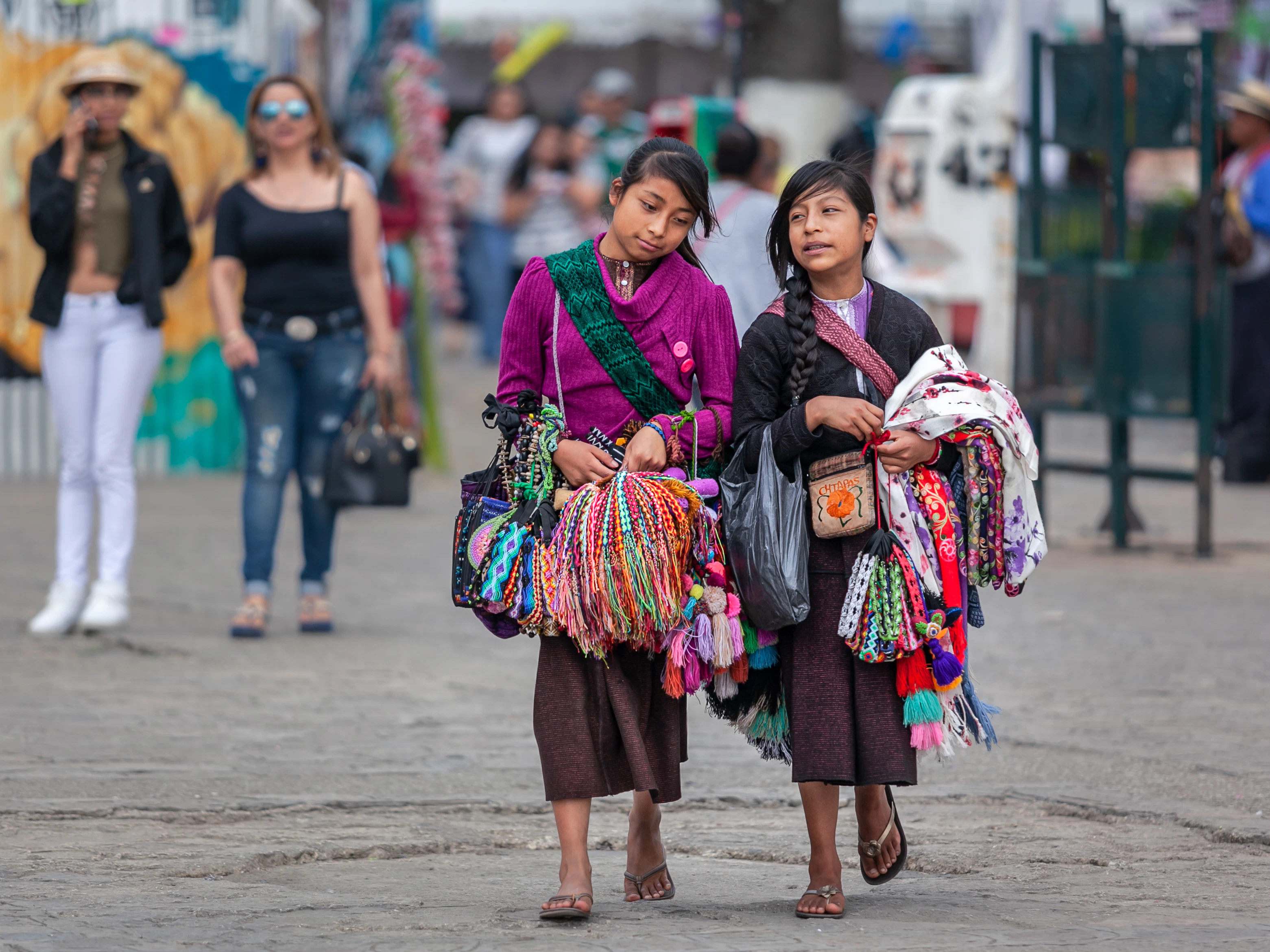 Two young girls in Indigenous outfits walk down a street with handicrafts to sell.