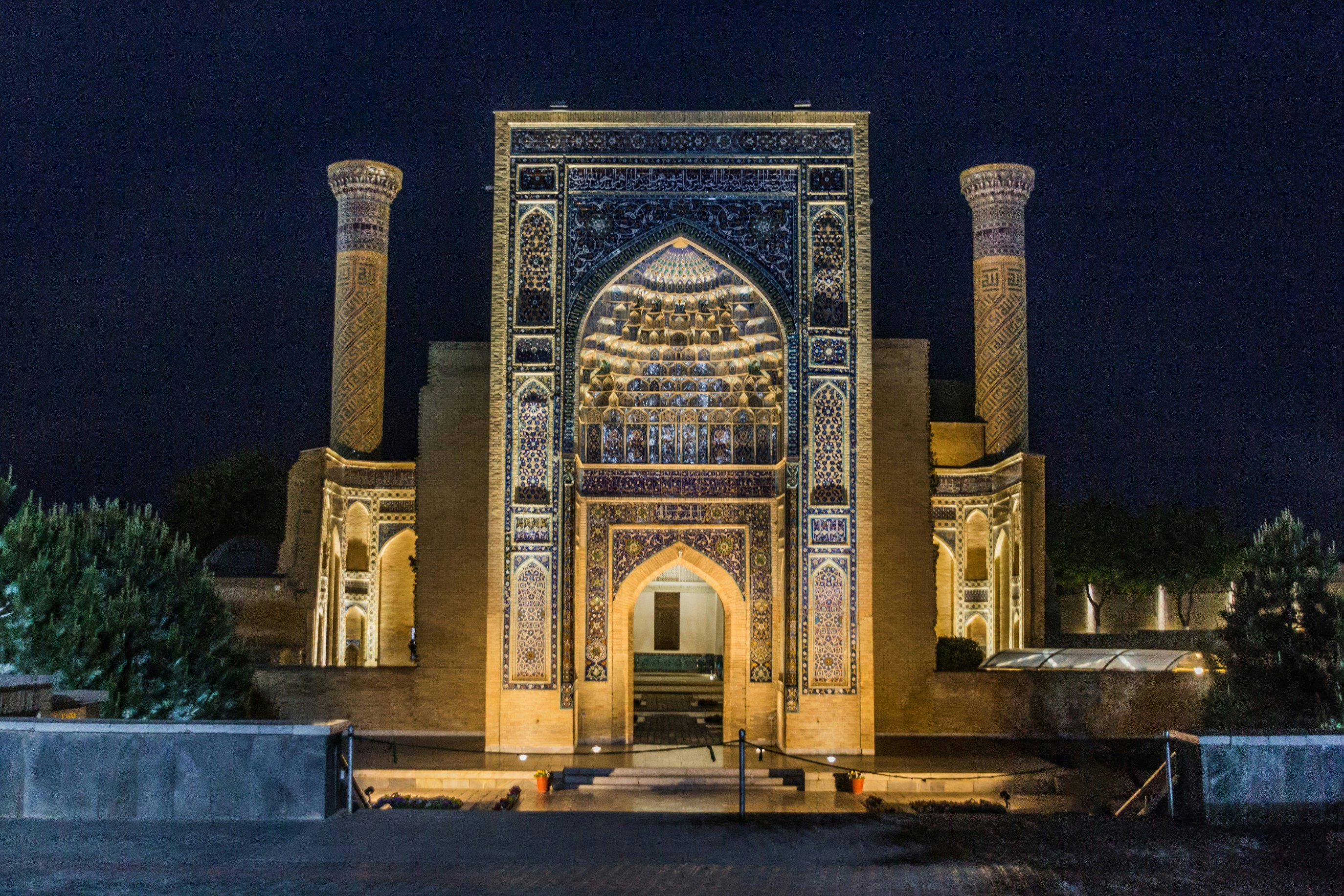 An evening view of the illuminated facade of a mausoleum, with blue tile in niches, Islamic arches and other details.