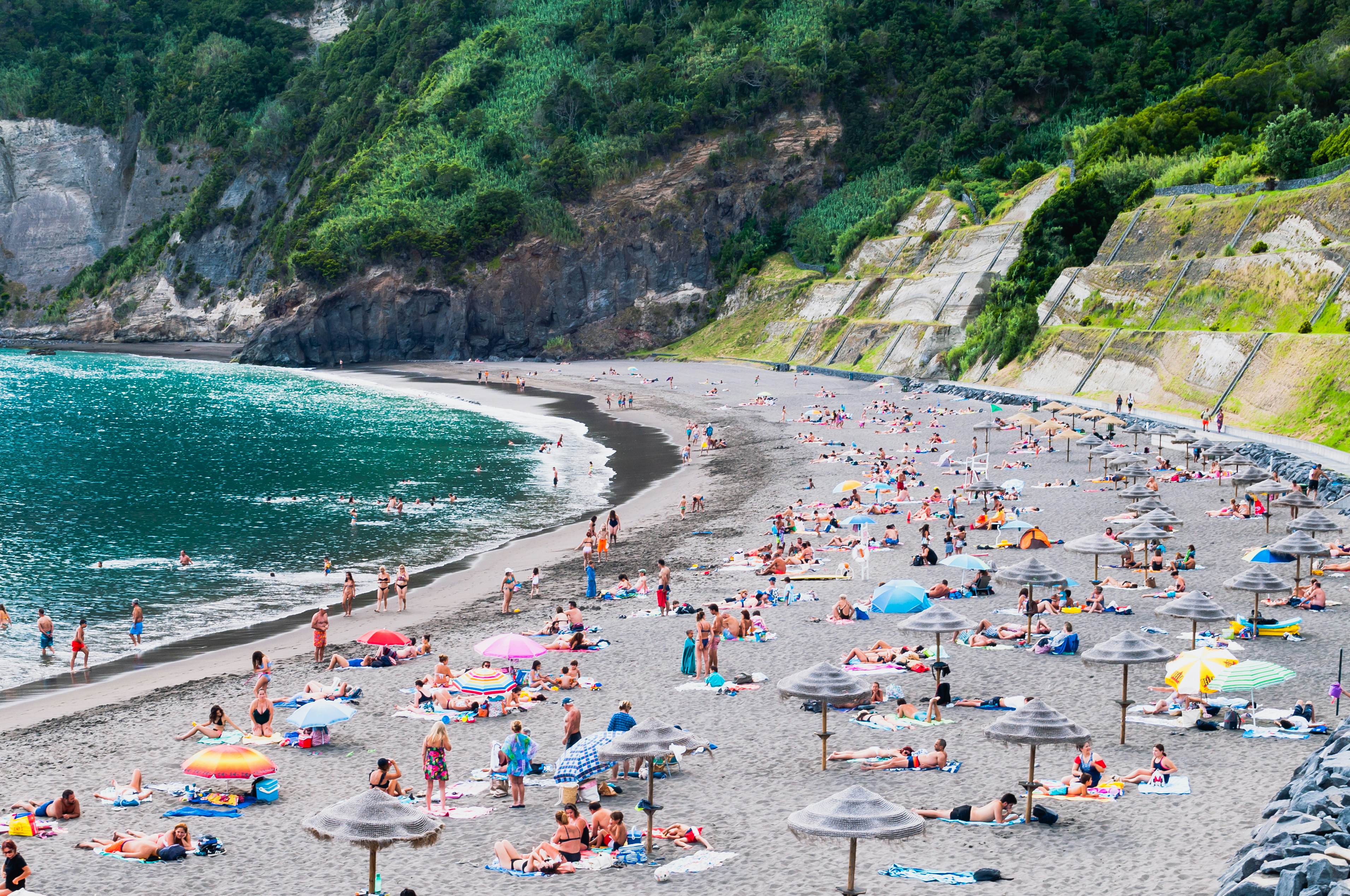 A wide shot of people in bathing suits on towels and under sun umbrellas on a black-sand beach. Cliffs covered with vegetation frame the cove of the beach.