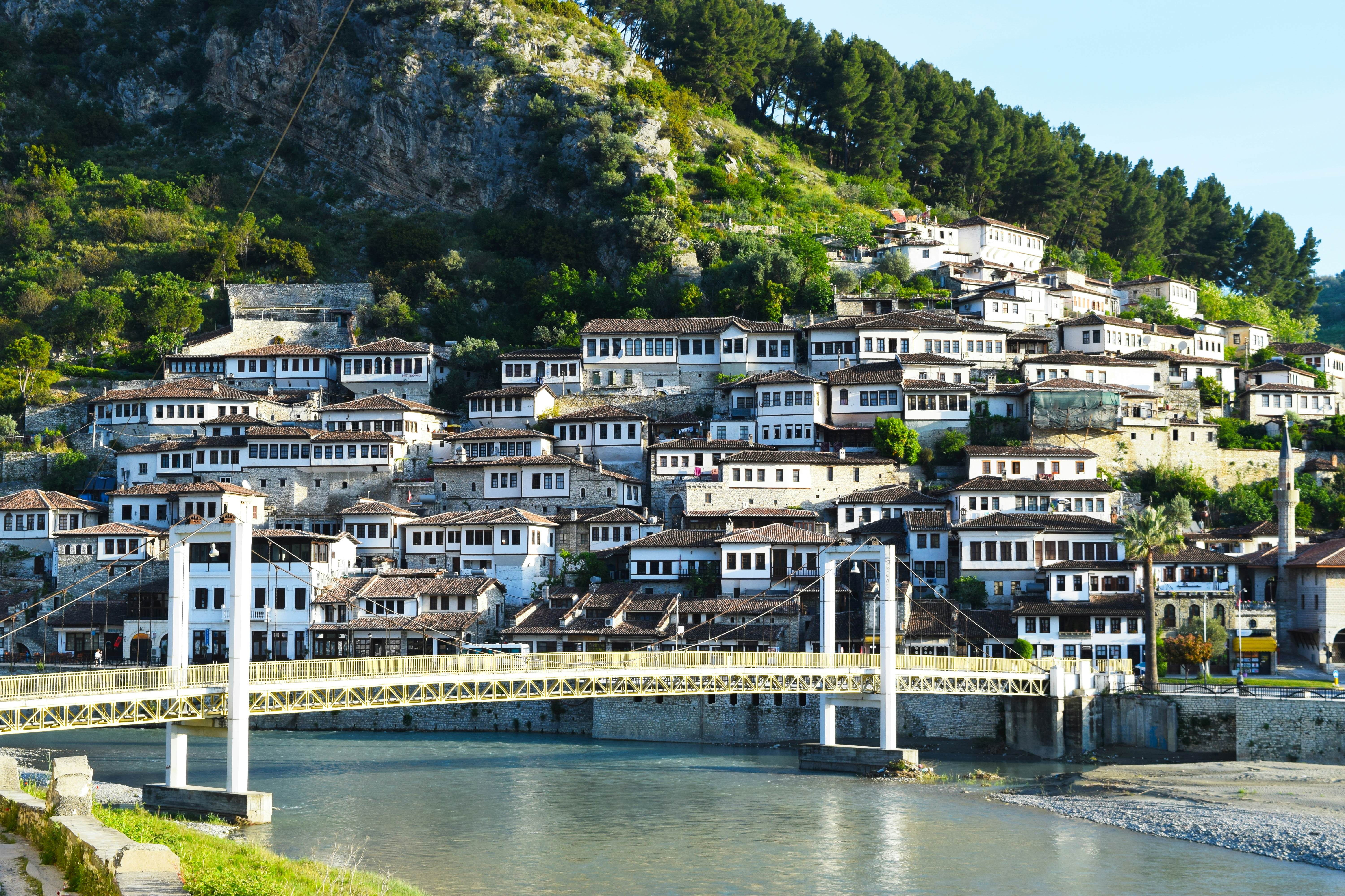 A small suspension bridge spans a river, with the white houses of a town rising on the hillside on the far bank.