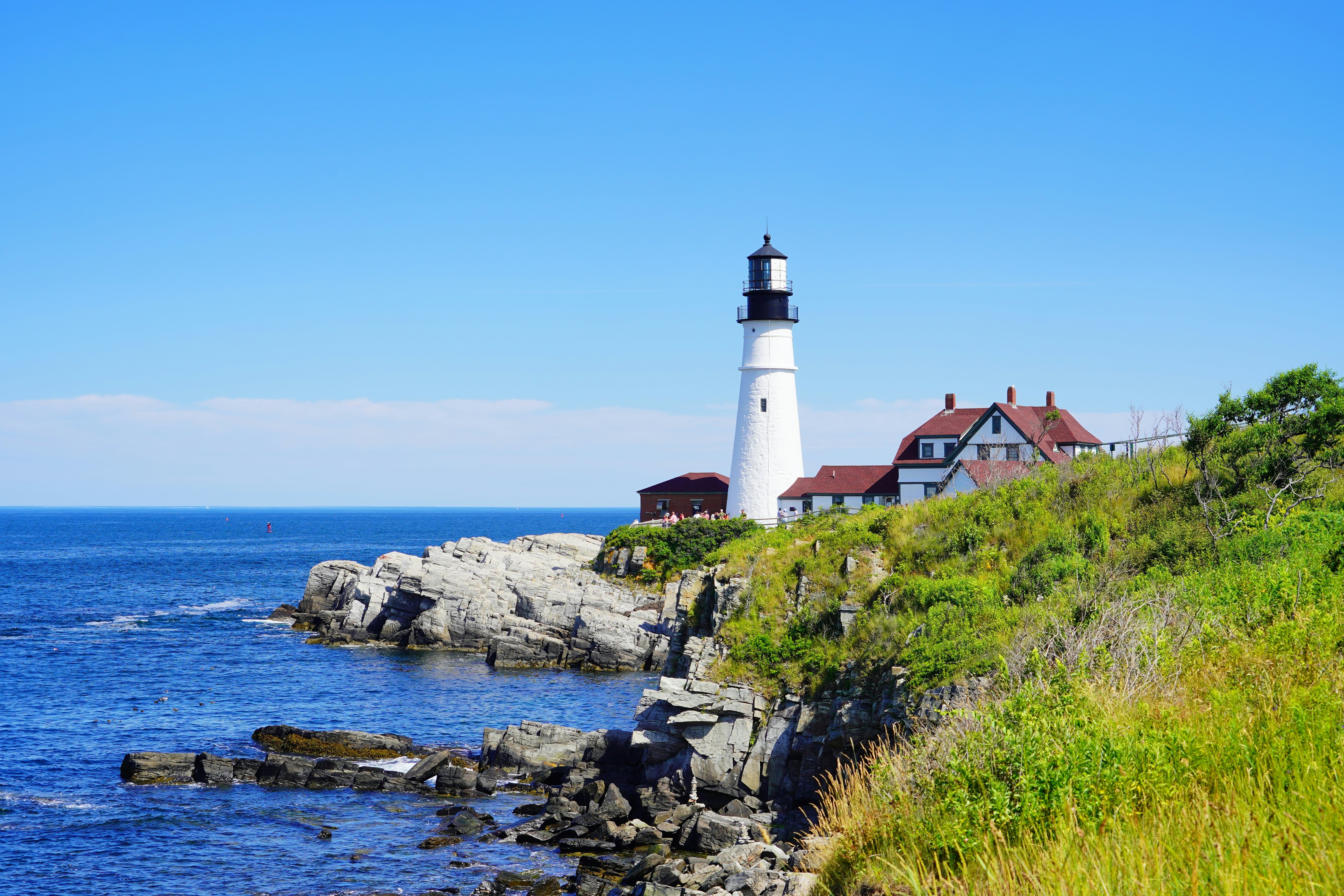 A tall lighthouse painted white stands at a rocky headland along the coast. A red-roofed house sits next to the lighthouse.