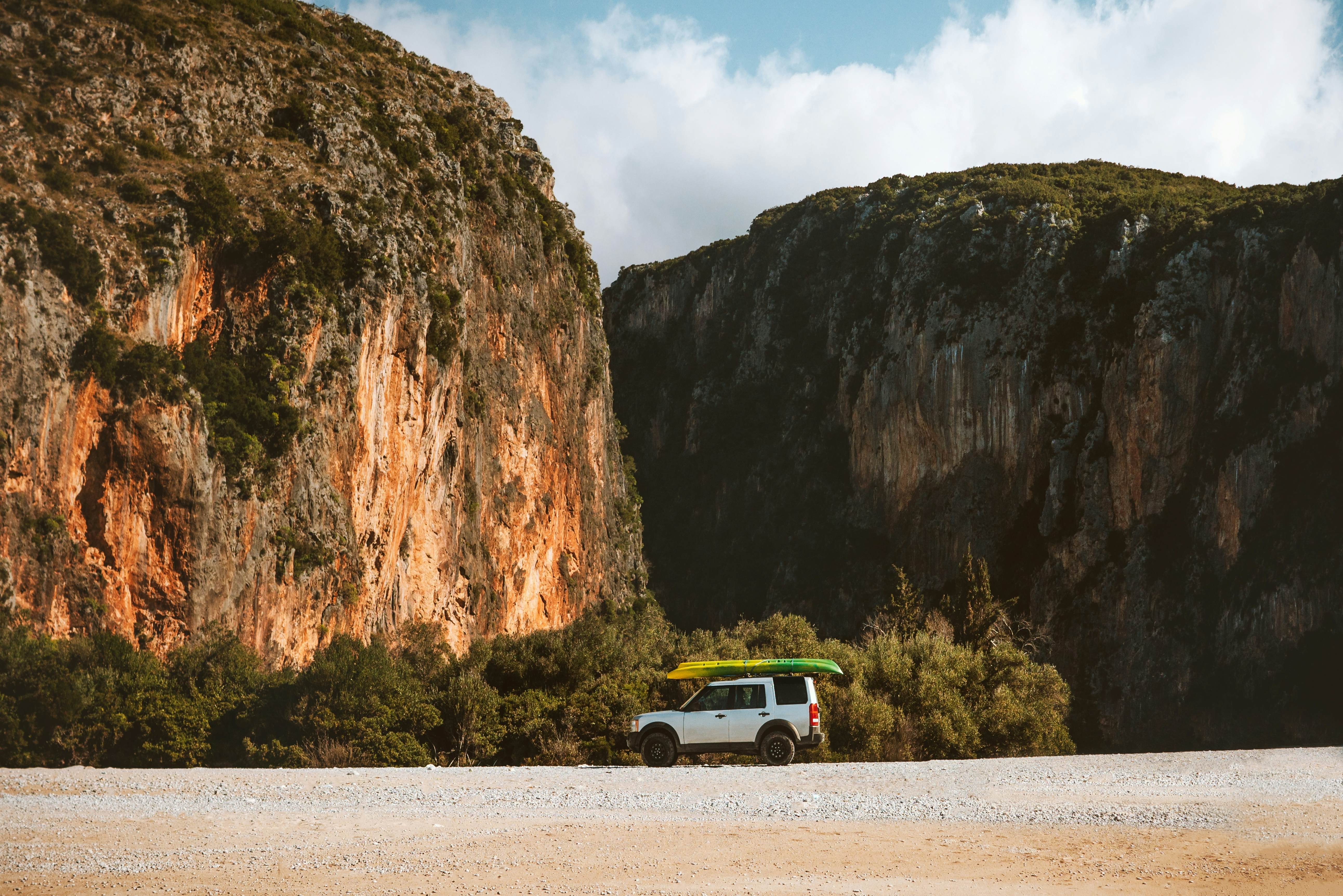 A van with a kayak on its roof is parked on a deserted beach. Dramatic cliffs rise directly behind the beach, forming a canyon.