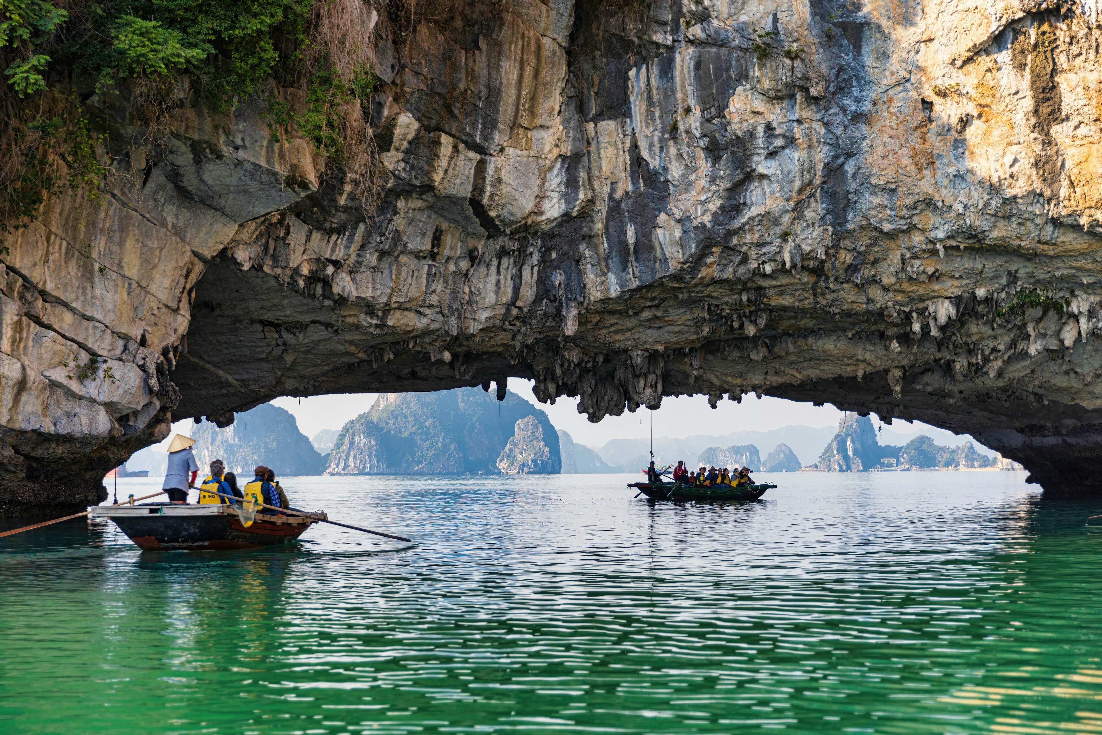 Two boats pass under an archway formed by rocks in a tropical bay with beautiful green water. Karst formations are visible in the distance, through the arch.