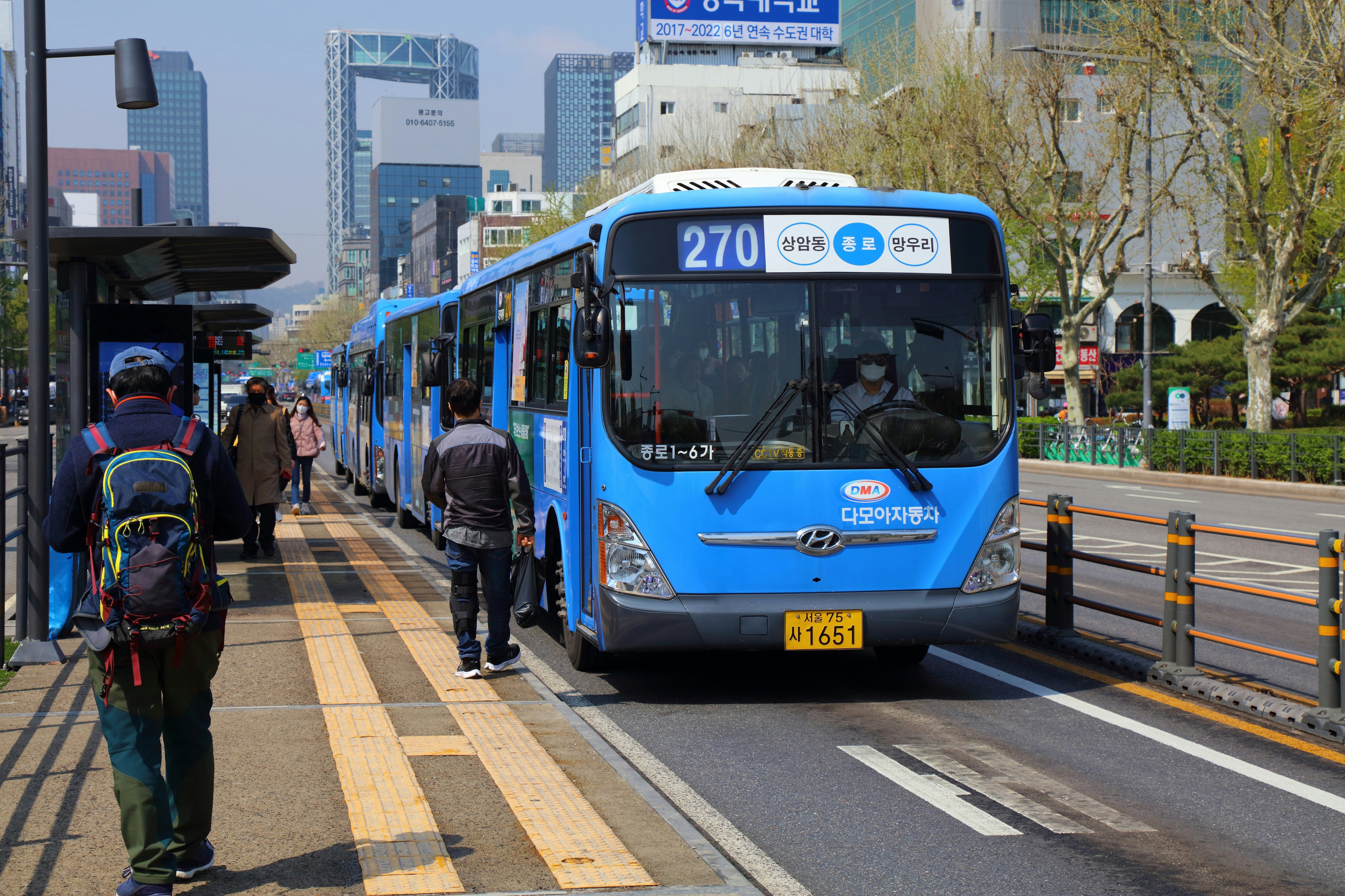 People board blue-painted buses in a dedicated bus lane on a wide city street. Office buildings are visible in the background.