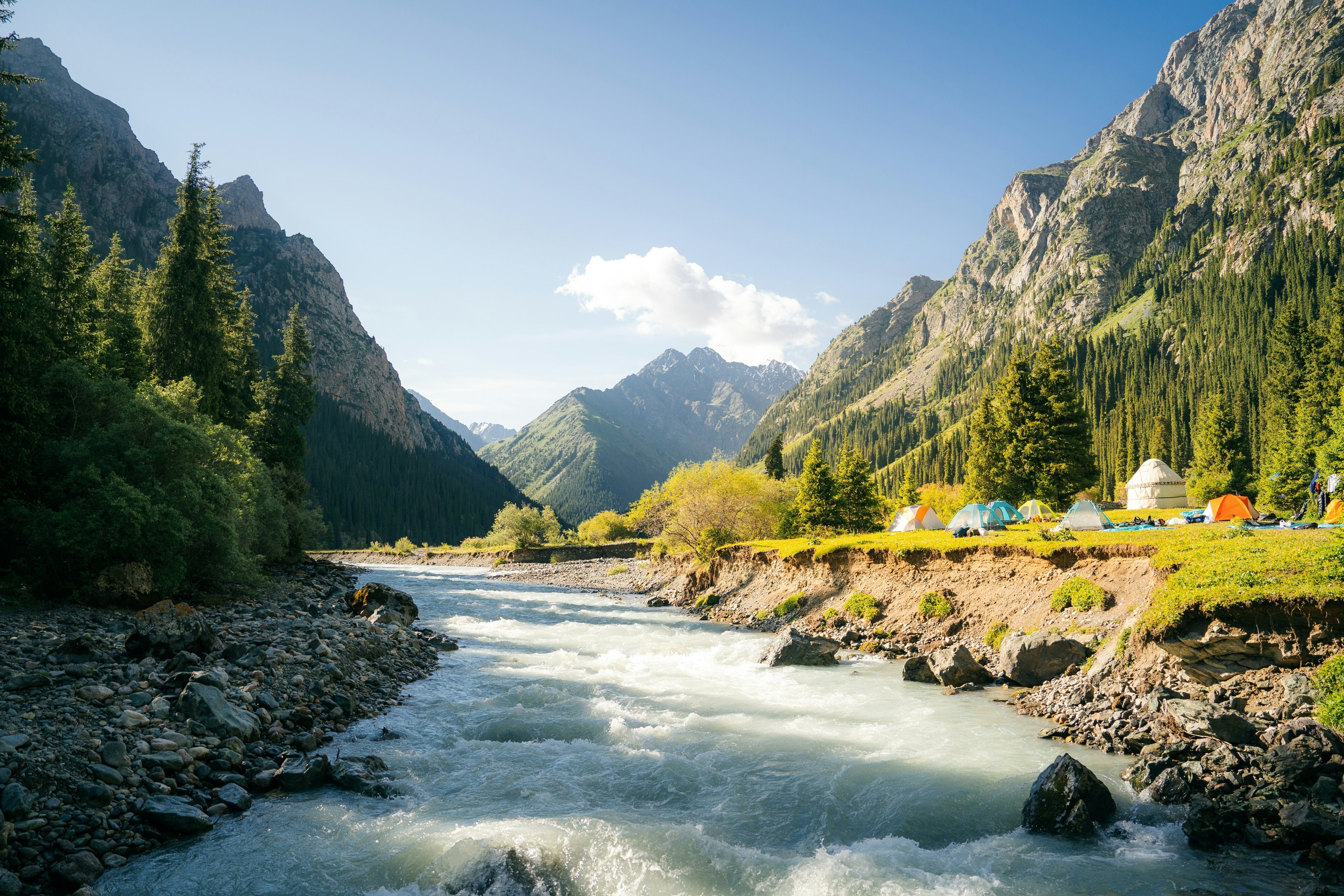 A wide gushing river flows through a mountain valley on a sunny day. Several colorful tents and a yurt stand in a nearby meadow.