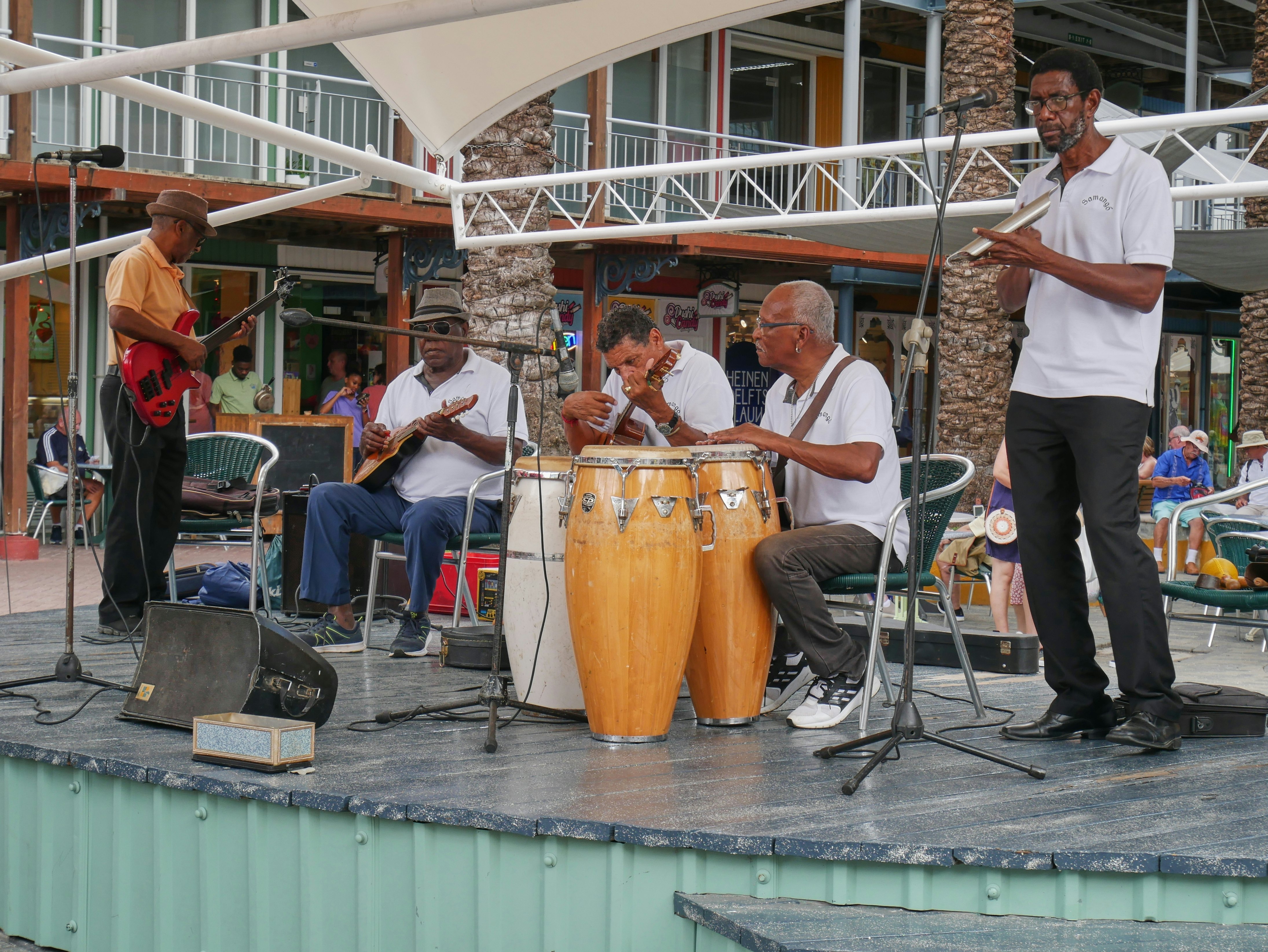 A band performs on a small stage in a city street. Three guitarists and two percussionists playing large drums and other instruments are pictured.