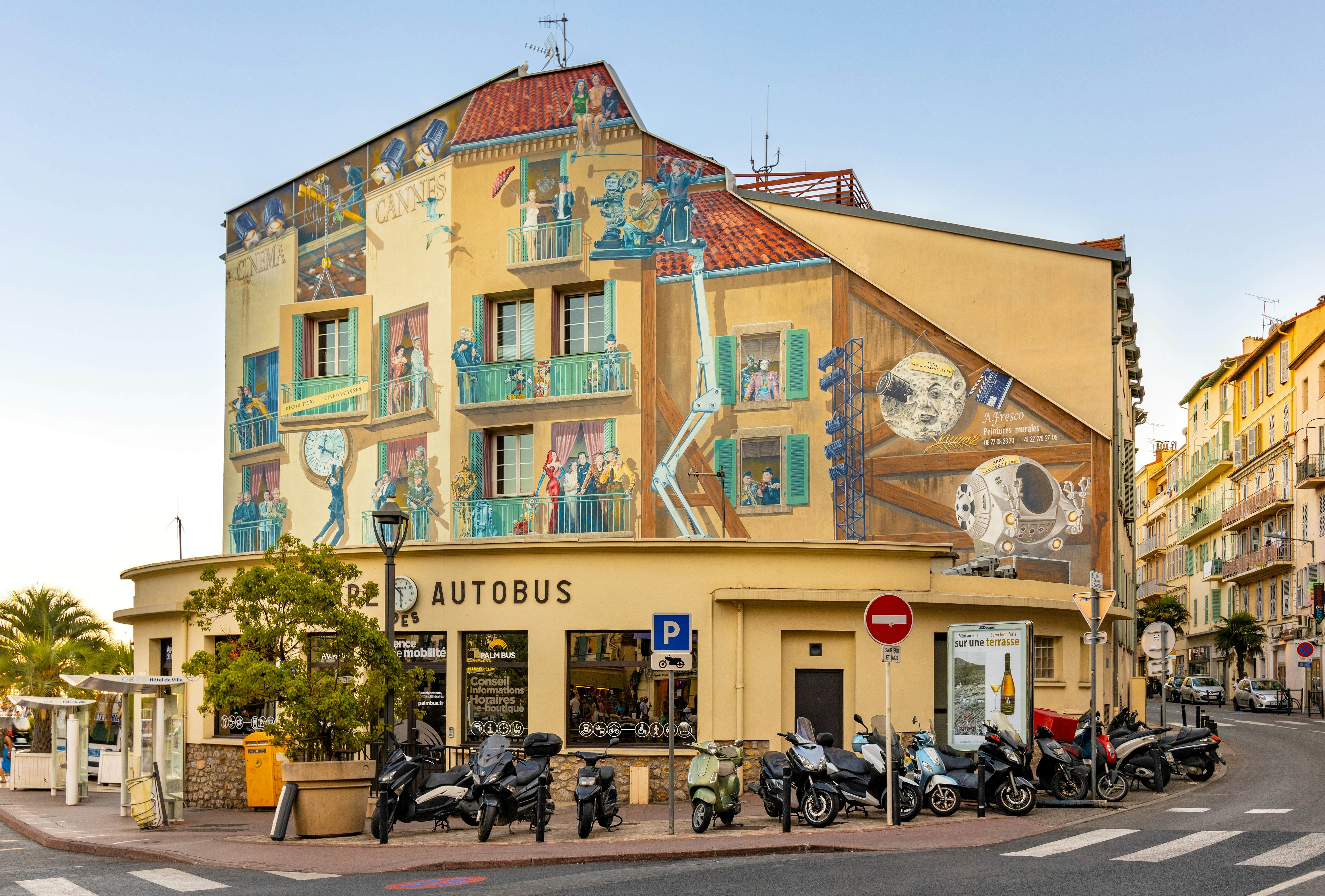 A yellow-colored building is adorned with a colorful mural on a city street. Motorcycles are parked in front of the building.