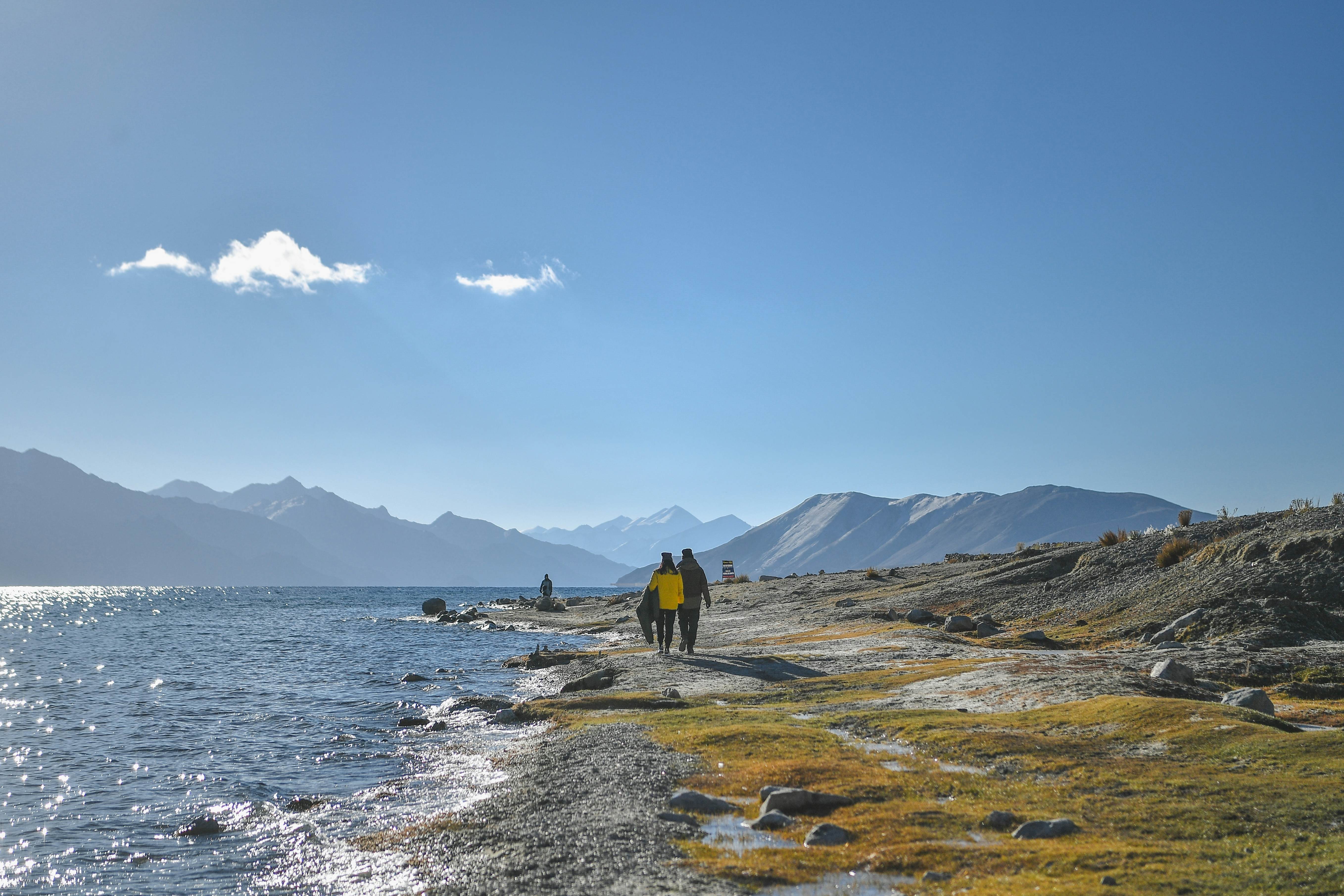 People walk along the coast of a mountain lake. Mountain peaks are visible in the far distance.