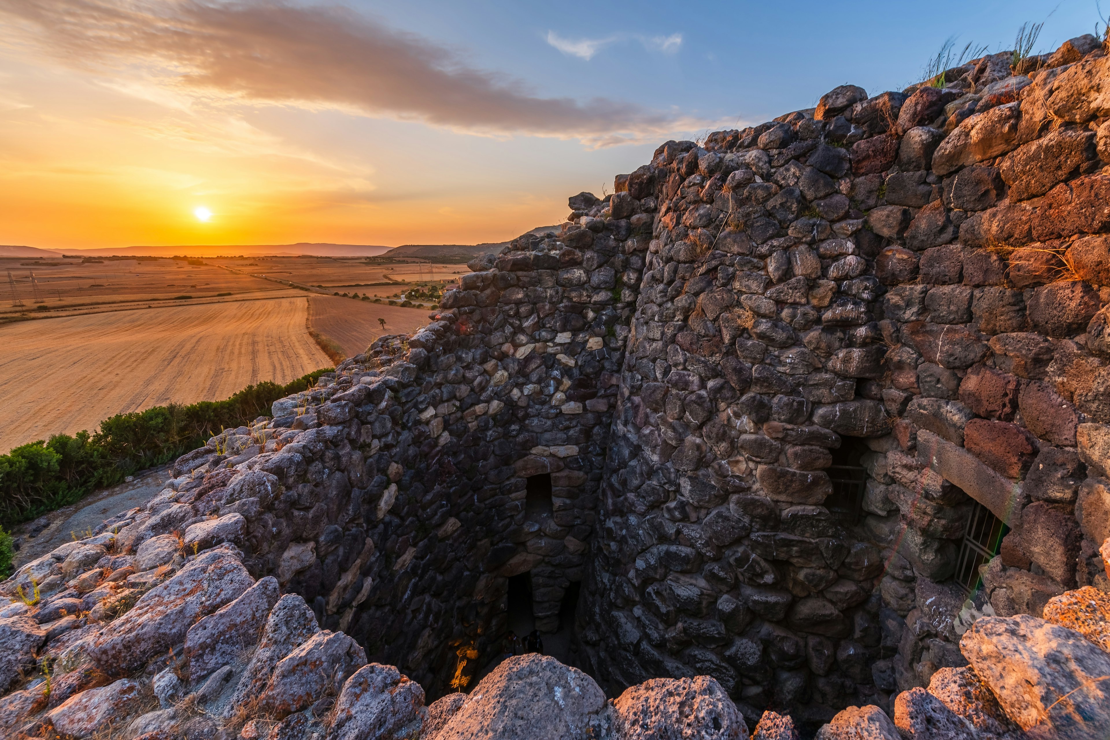 An ancient structure made of stones and mortar with windows and a pit is pictured at sunset. Fields are visible in the distance, lit by the setting sun.