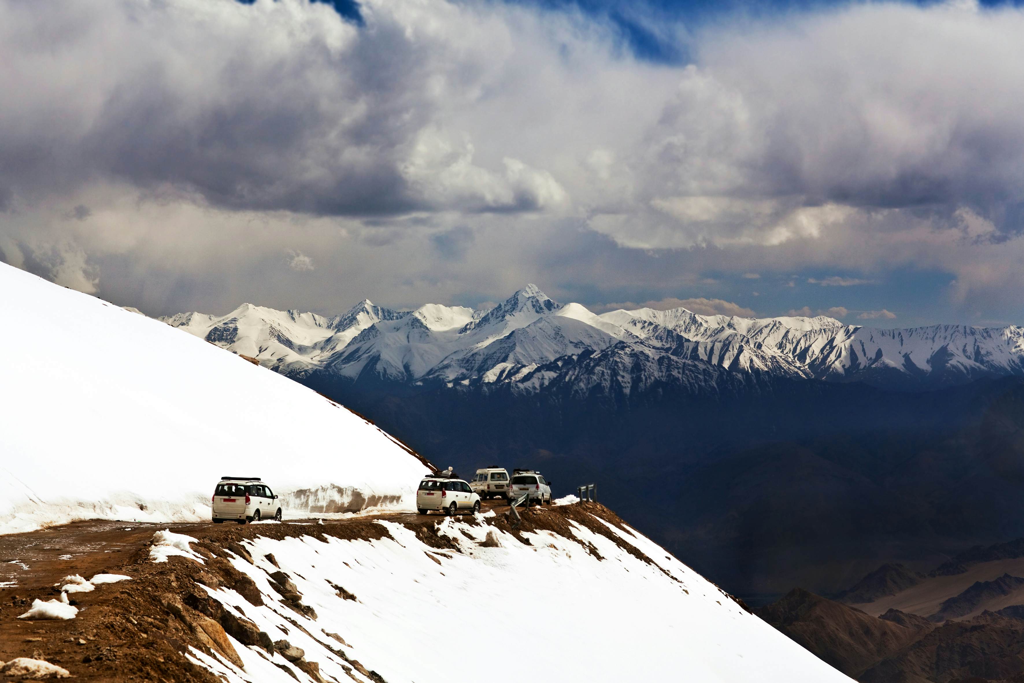 A row of vans follows a narrow road on a mountain pass, with slopes covered in snow. More snow-capped peaks are visible in the distance across a valley.