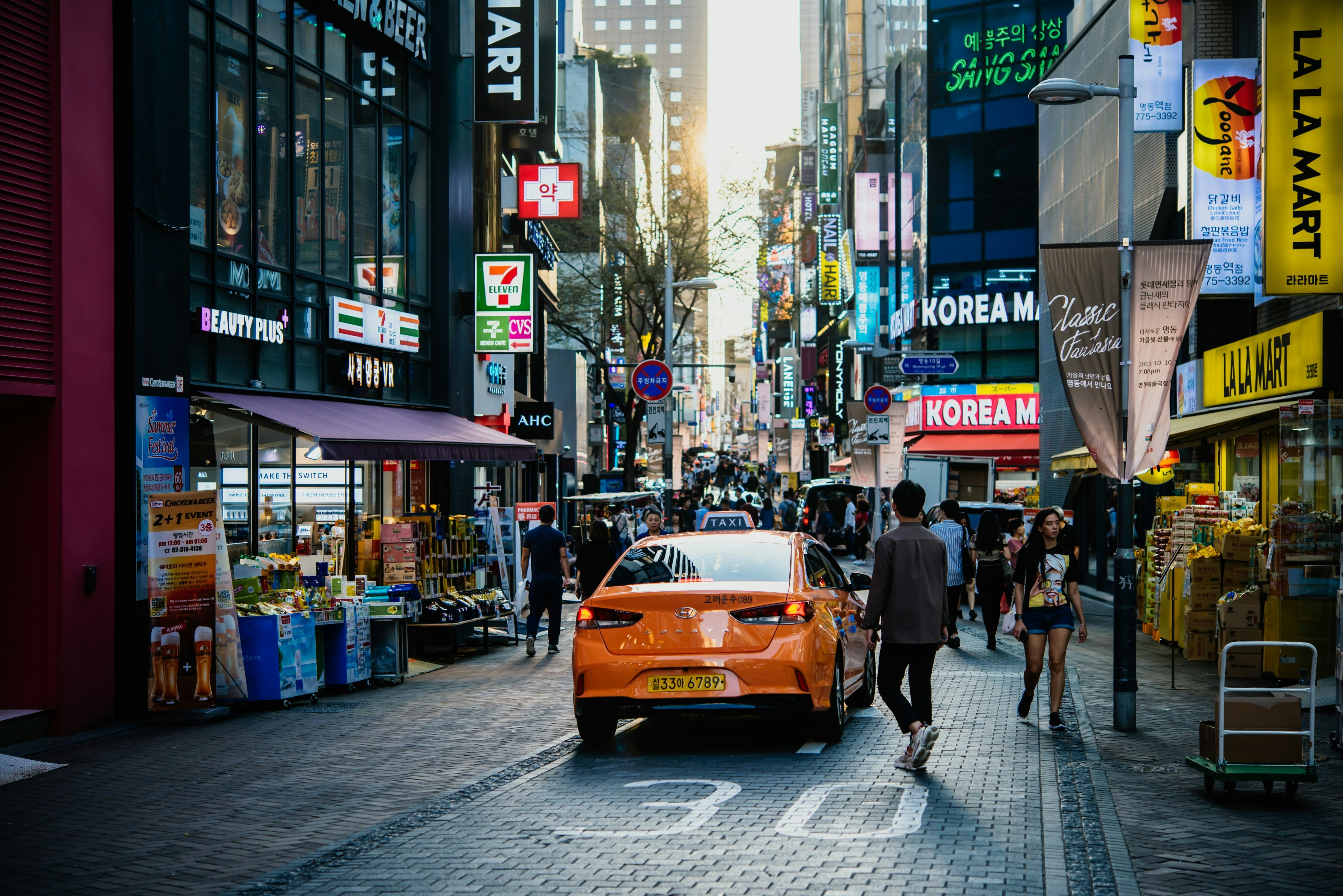 An orange taxi drives into a narrow street crowded with pedestrians and businesses in a large city.