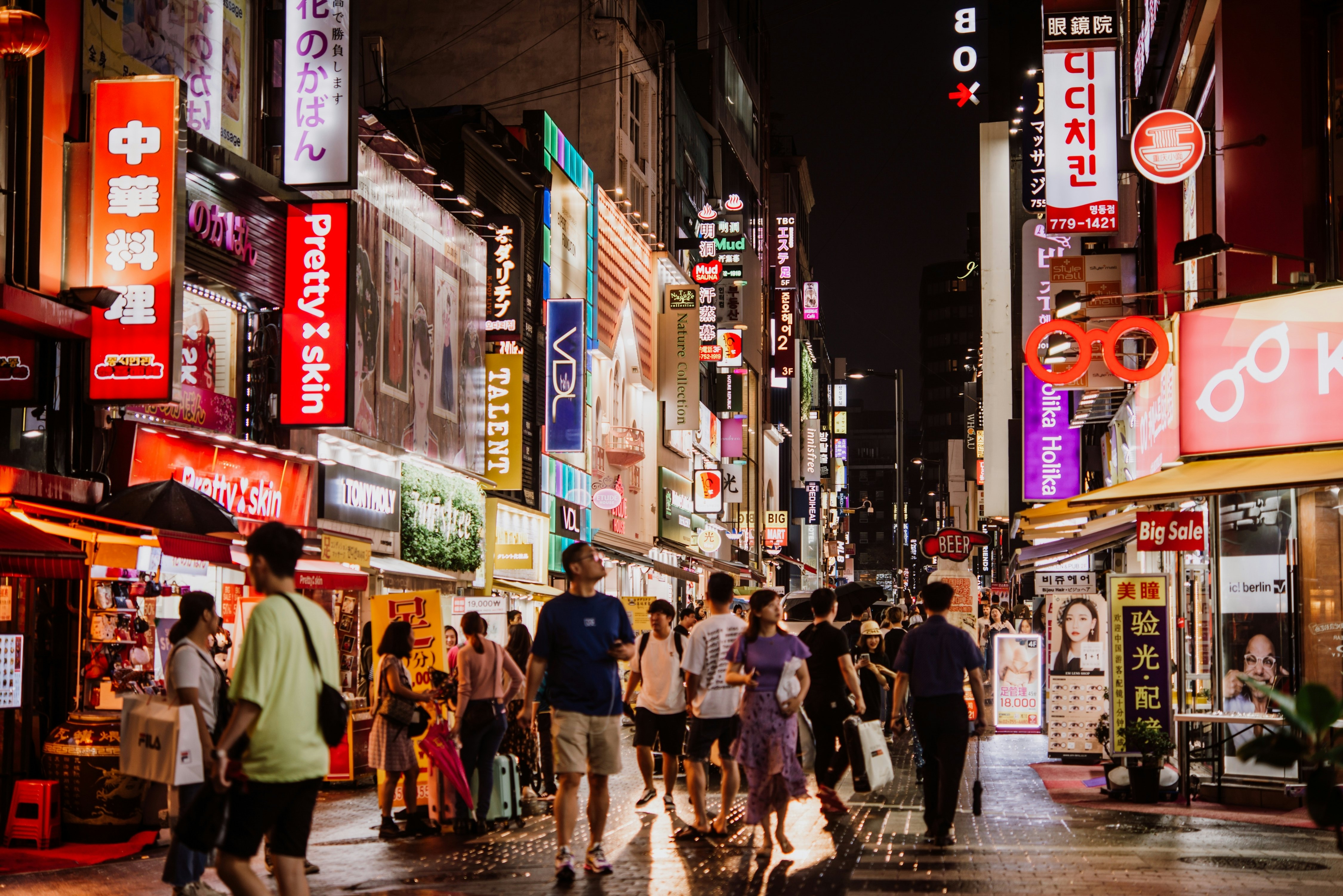 A crowd of people walk on pedestrian-only street at night in a large city. Lit-up signs and billboards flood the street scene with light.